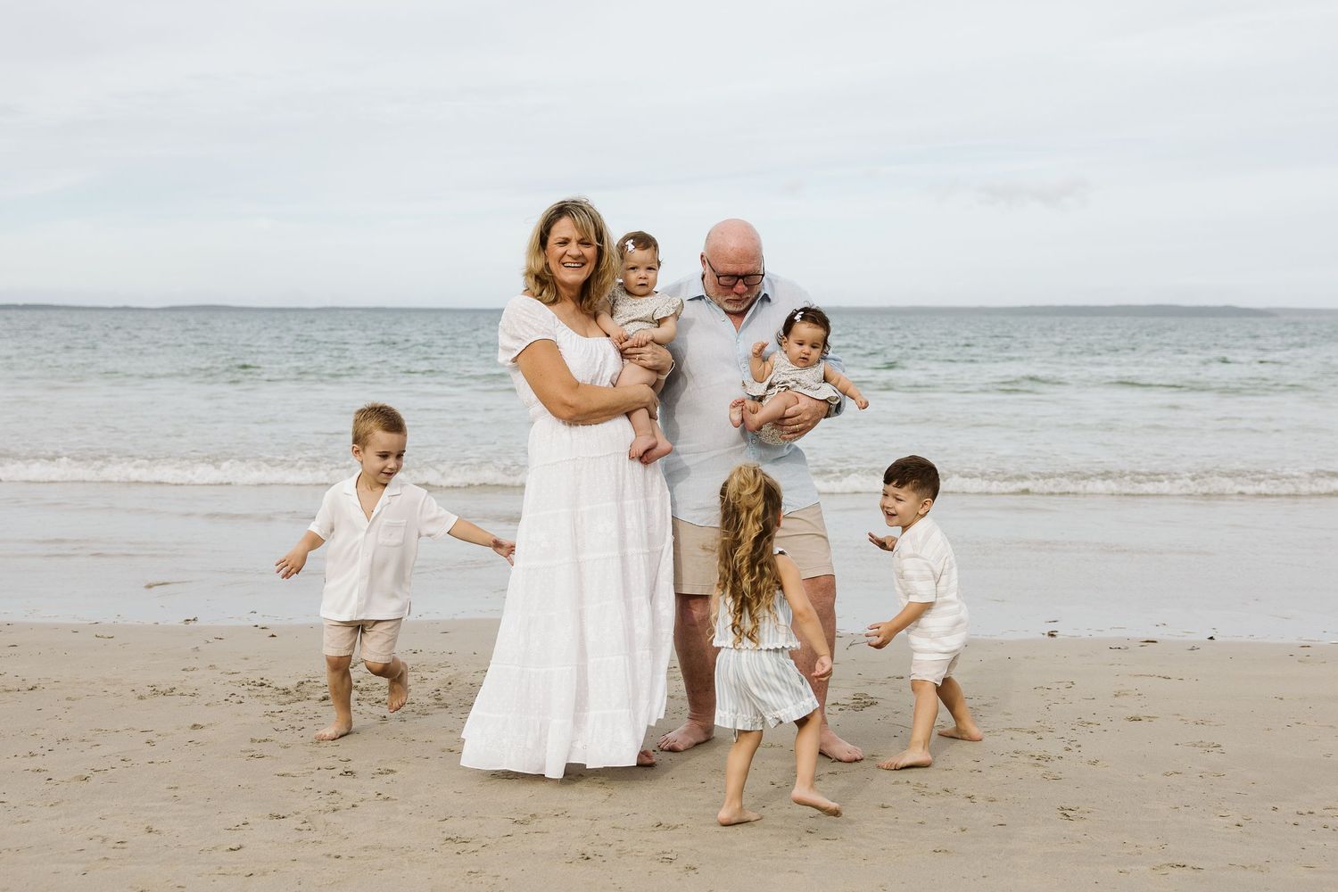Family of seven poses on a sandy beach, parents holding babies while children play around them in white outfits.