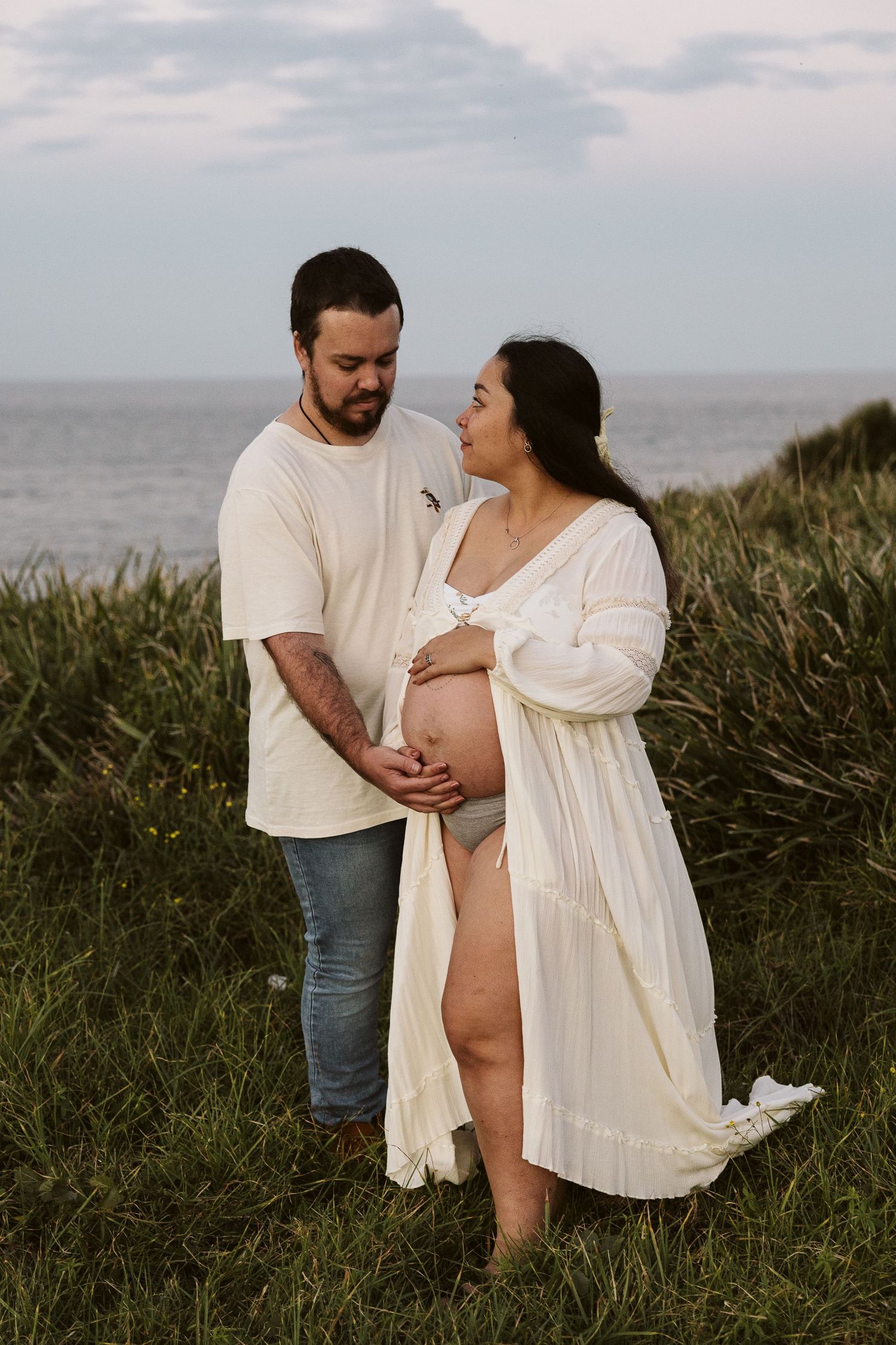Couple tenderly holding pregnant belly outdoors near ocean grass, woman in white robe, man in cream shirt and jeans.