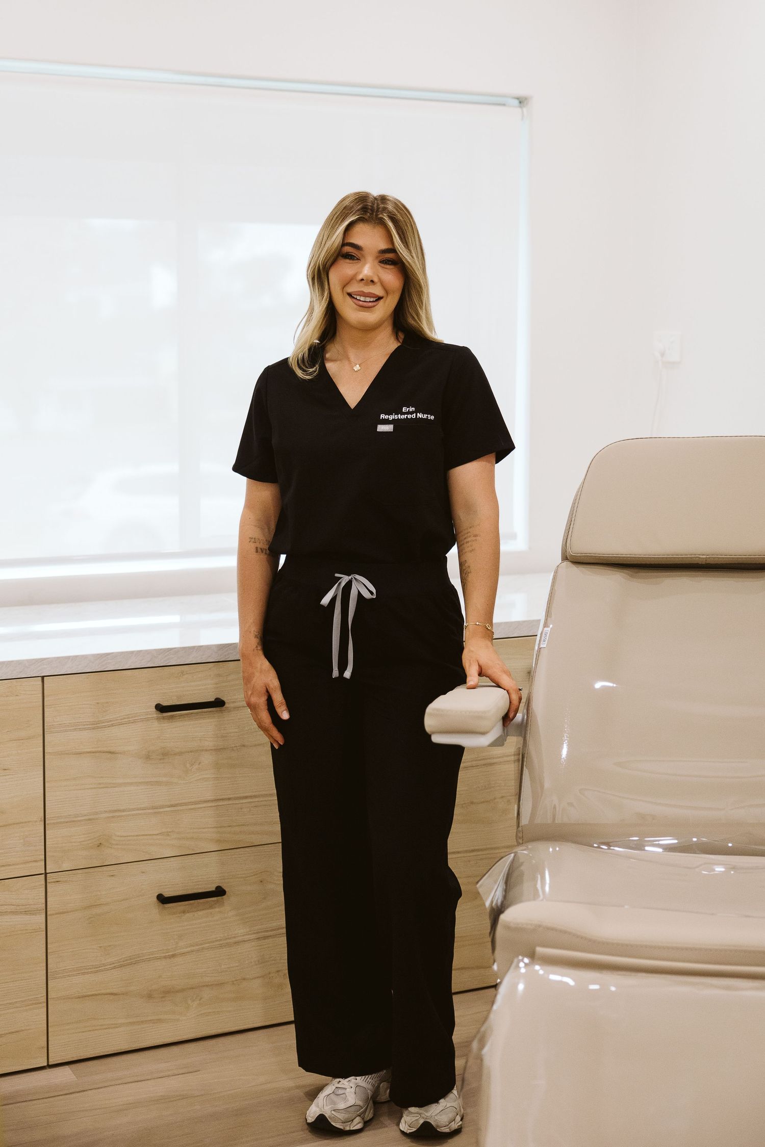 Female skincare professional in black scrubs standing beside treatment chair in a modern aesthetics clinic room.