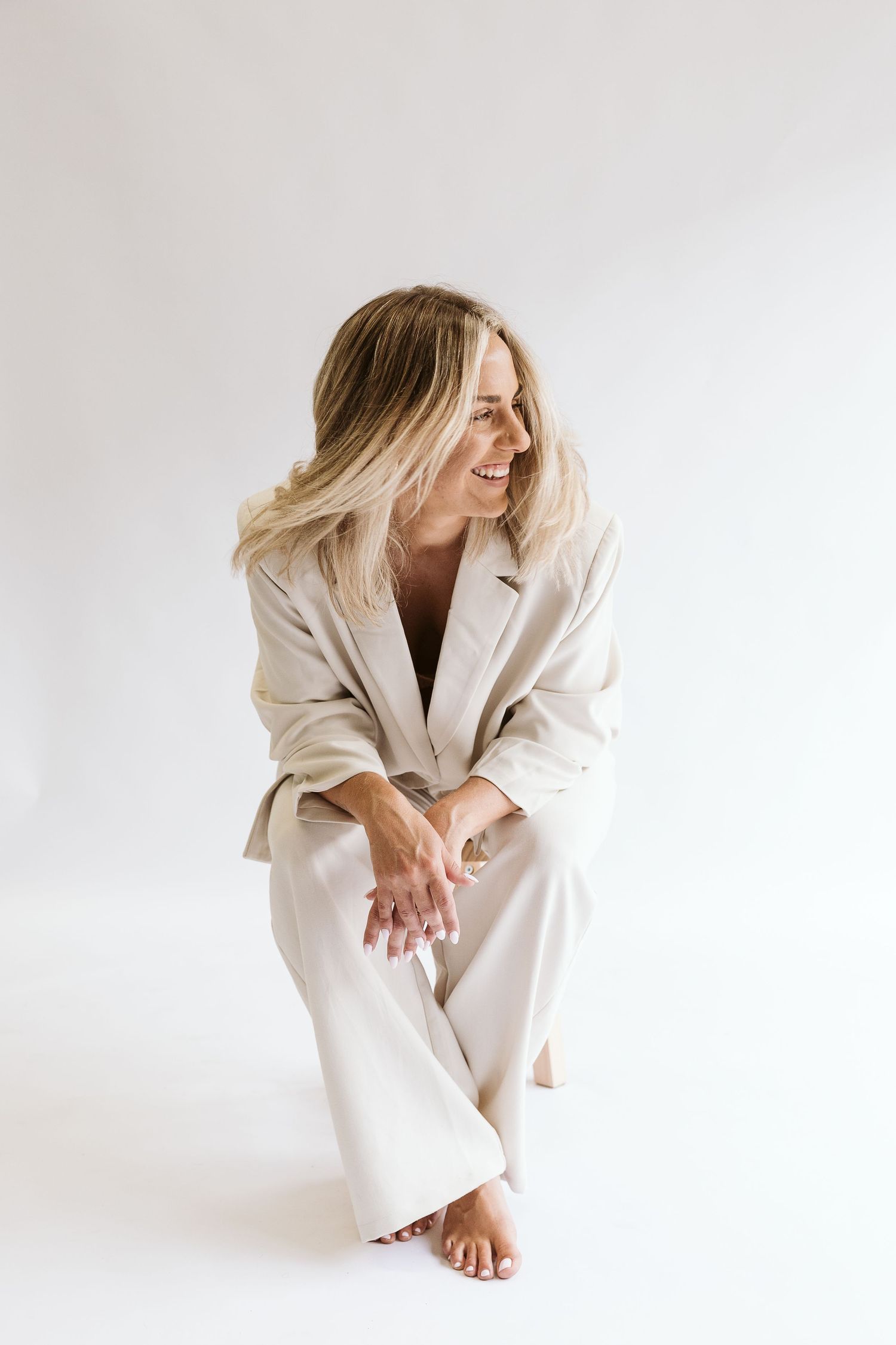 Laughing blonde woman in cream blazer and trousers crouches on white background.