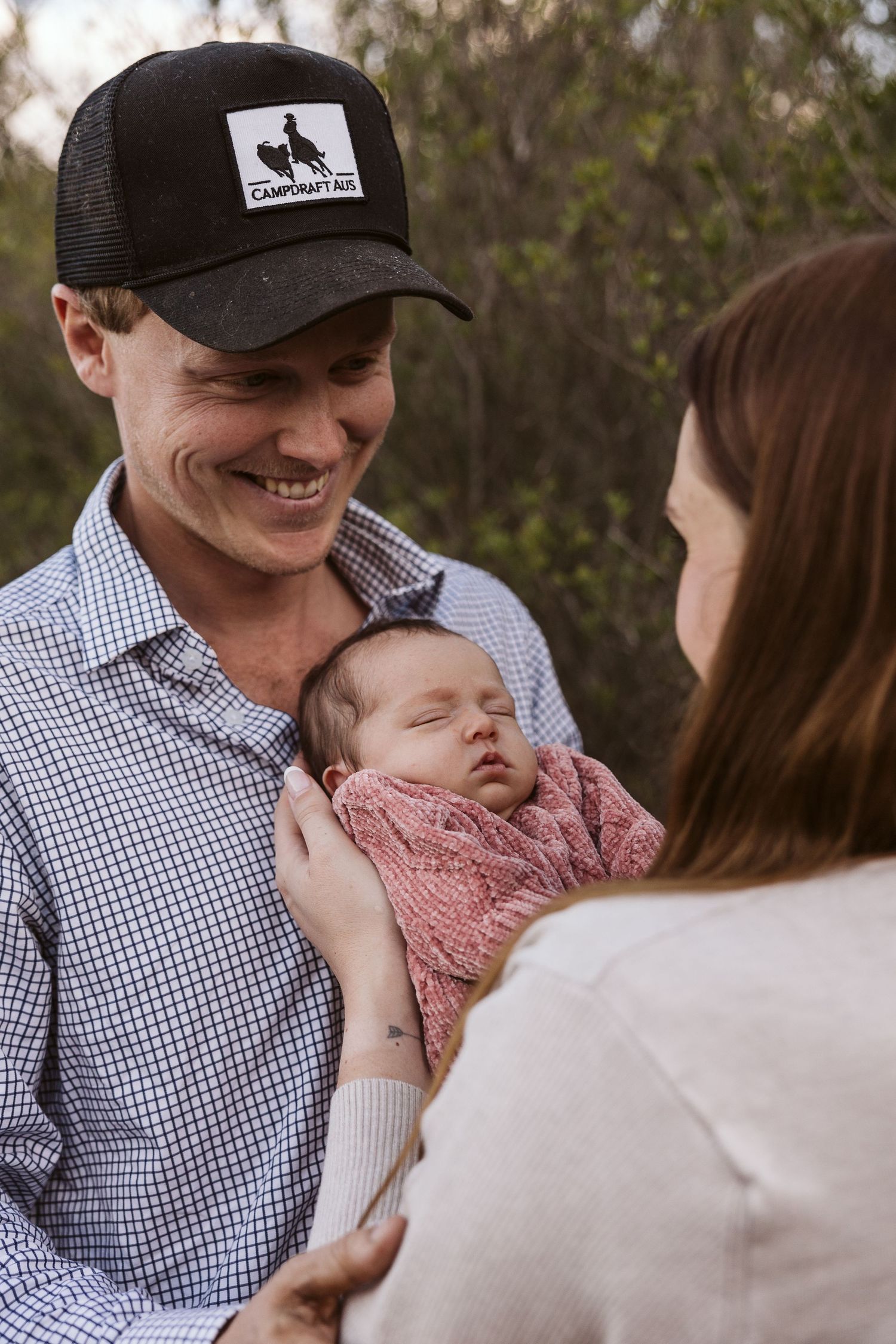 Smiling father in black cap holding sleeping newborn while mother looks on lovingly.