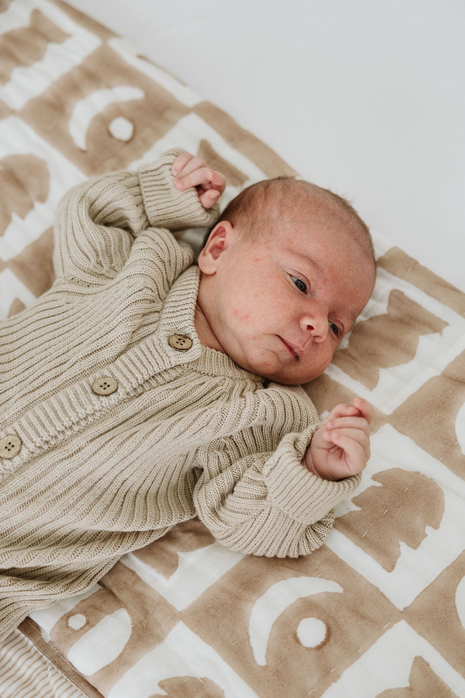 Newborn baby in beige knit cardigan lying awake on a patterned cream and tan blanket.