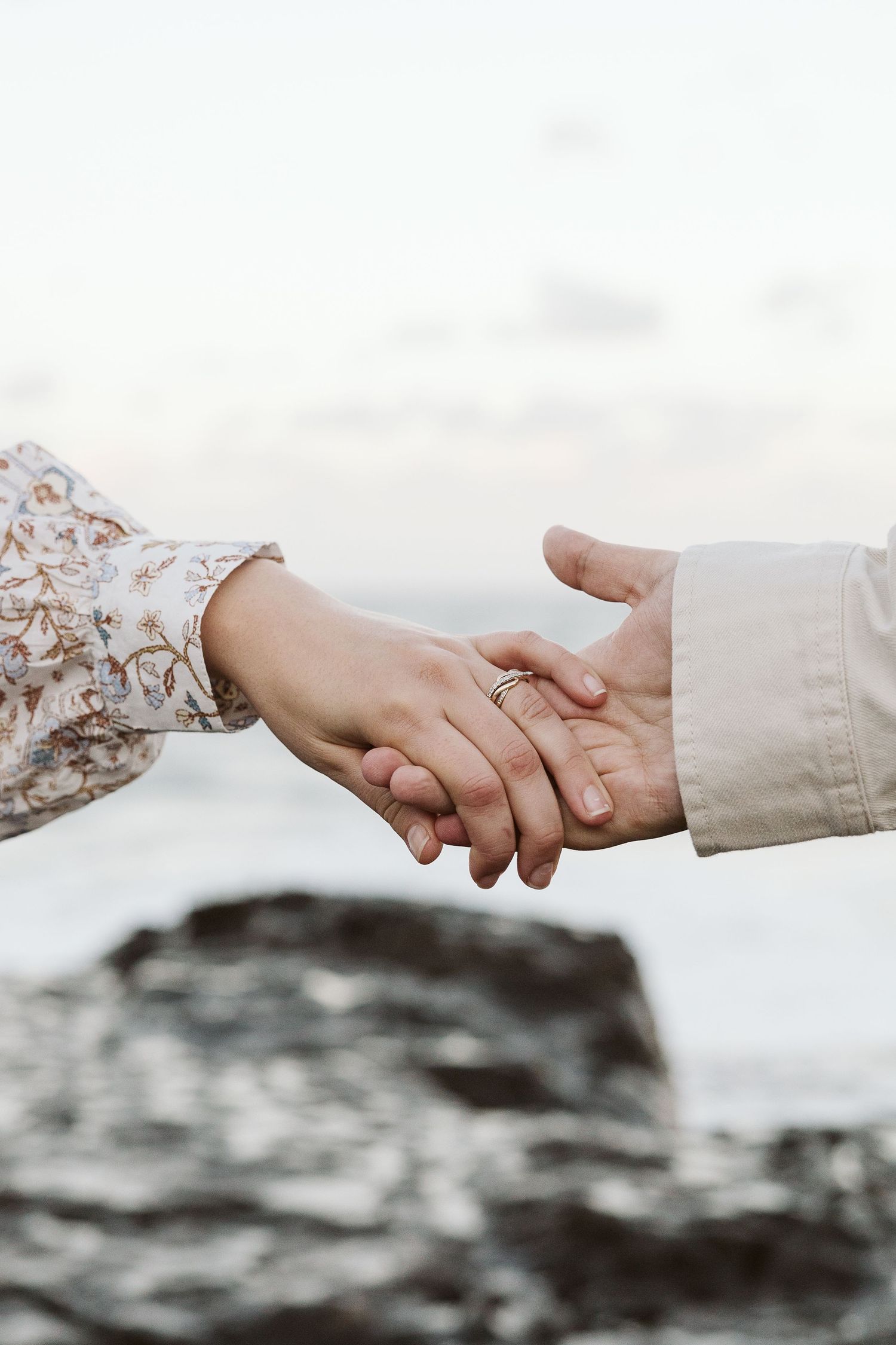 Close-up of couple holding hands with engagement ring visible, rocky coastal backdrop at dusk.