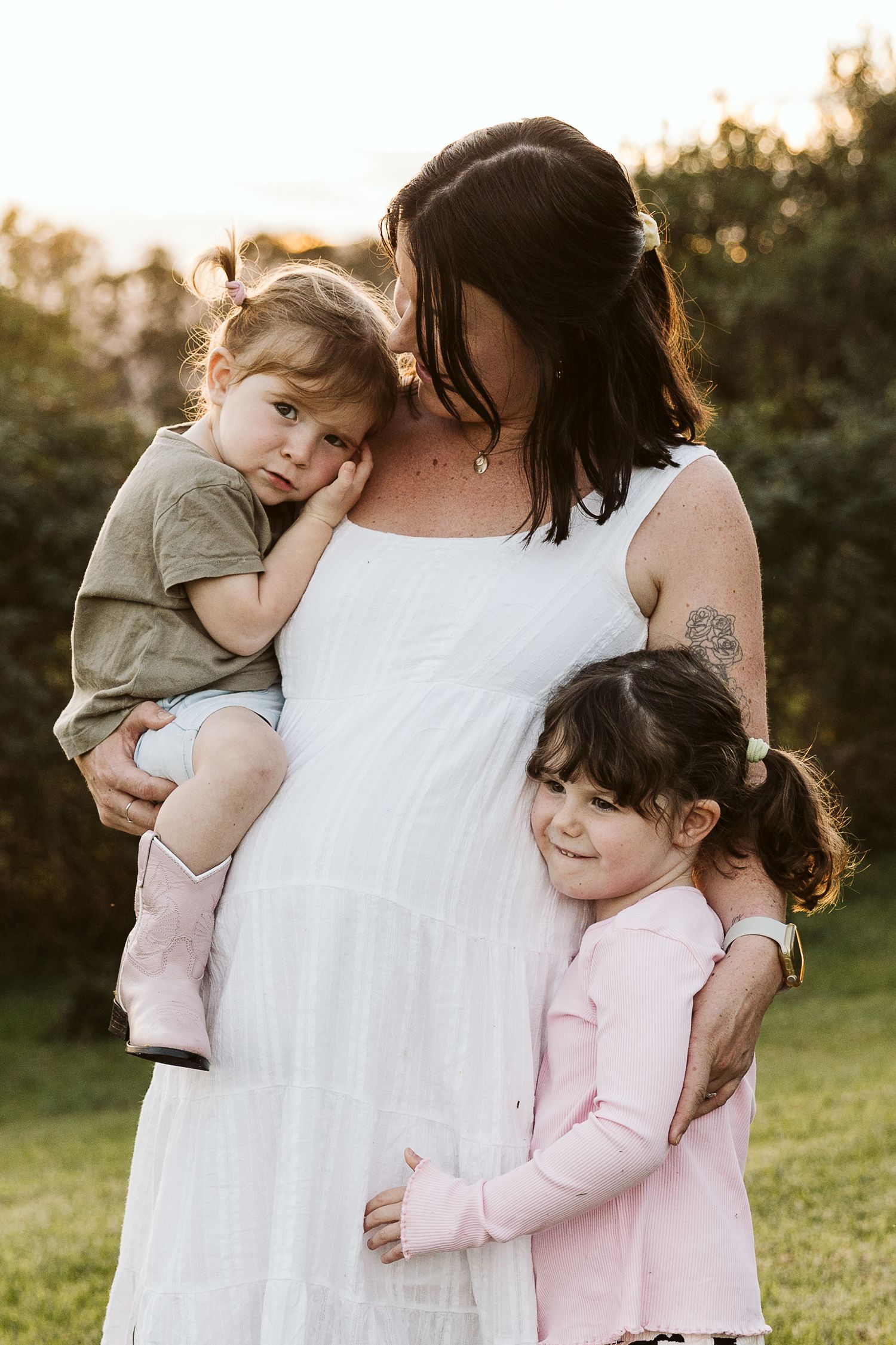 Pregnant mother in white dress holding toddler while older daughter in pink hugs her at golden hour outdoors.