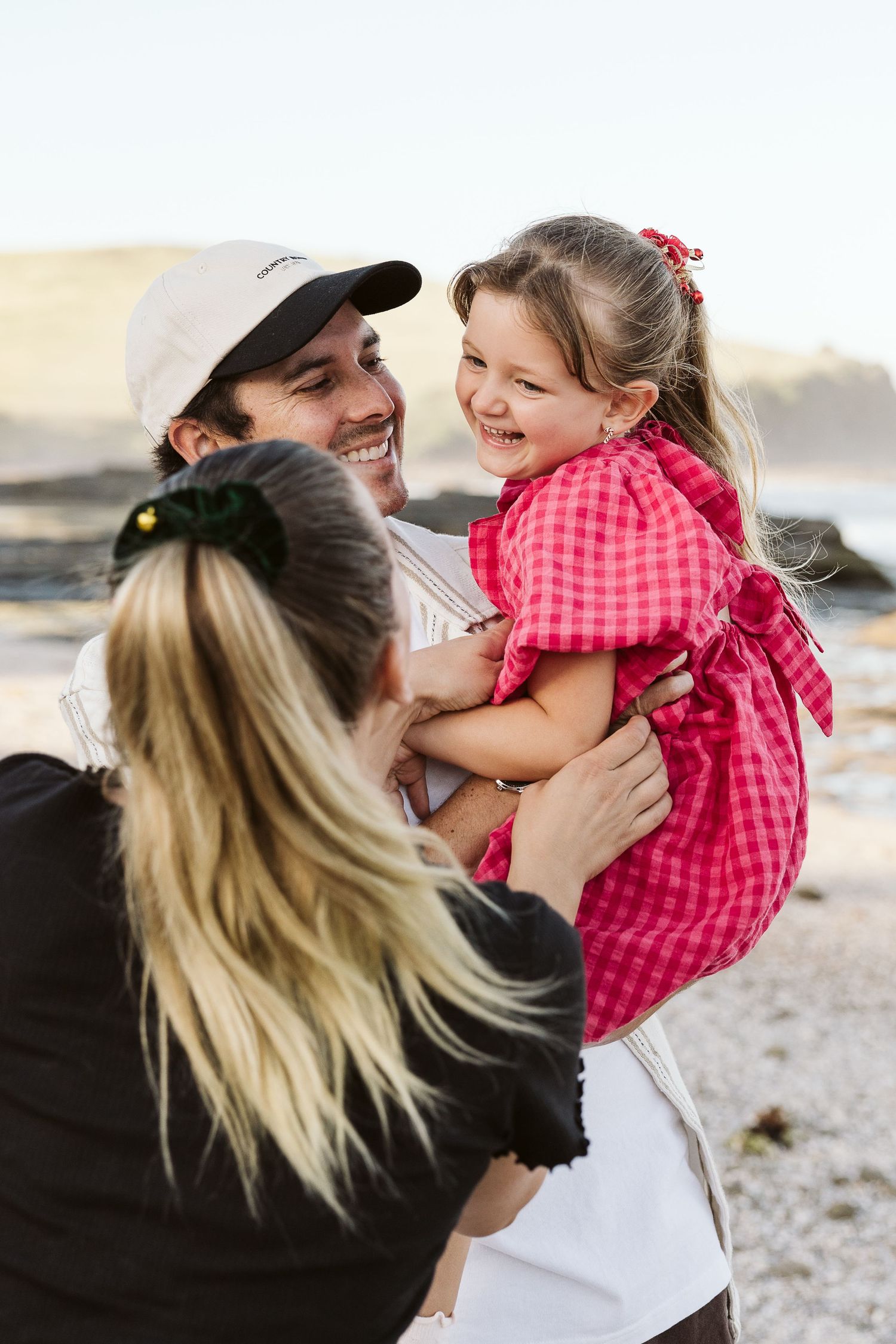 A happy family at the beach, with a man in a white cap holding a laughing girl in a red dress while a woman looks on.