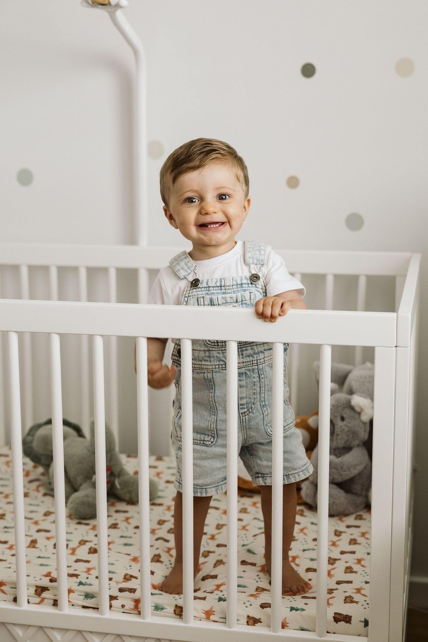 Smiling toddler boy in denim overalls standing in a white crib with stuffed animals in a nursery.