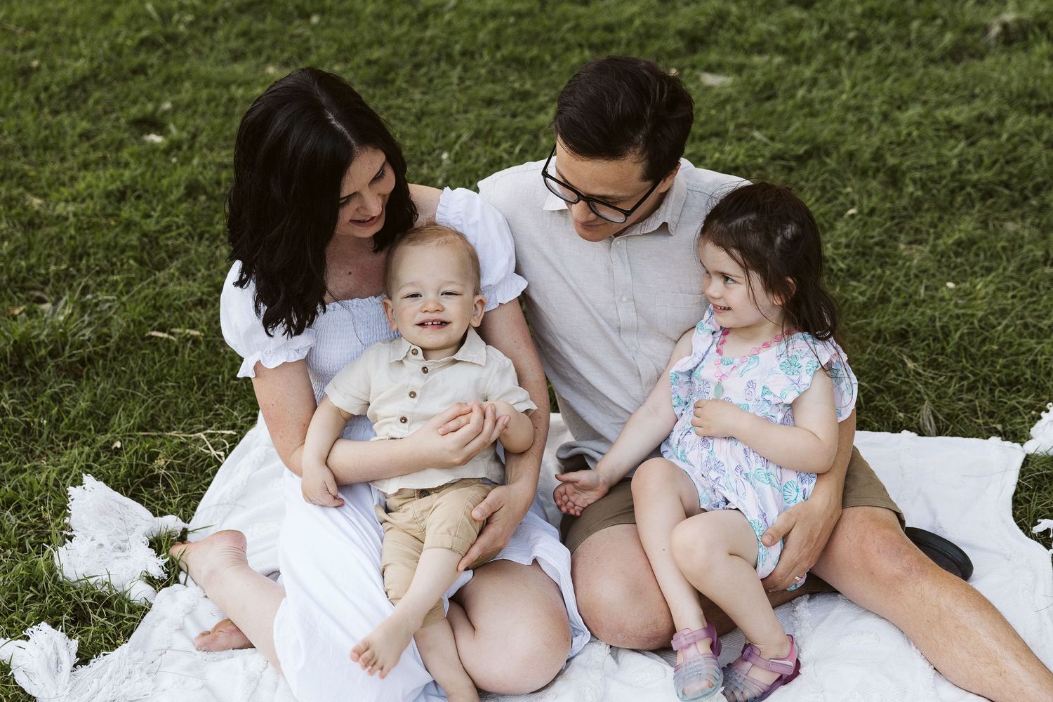Happy family of four sitting on a white blanket in a green park, parents with toddler boy and young girl smiling.