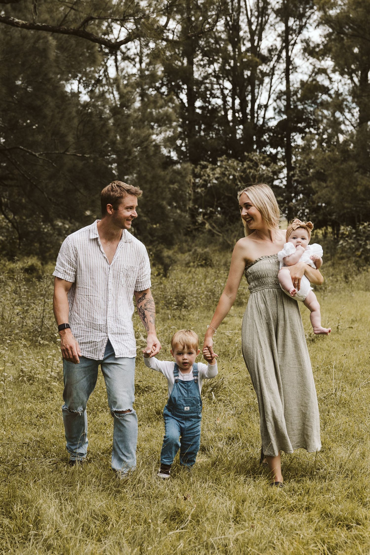 Family of four walking through green field holding hands with baby and toddler.