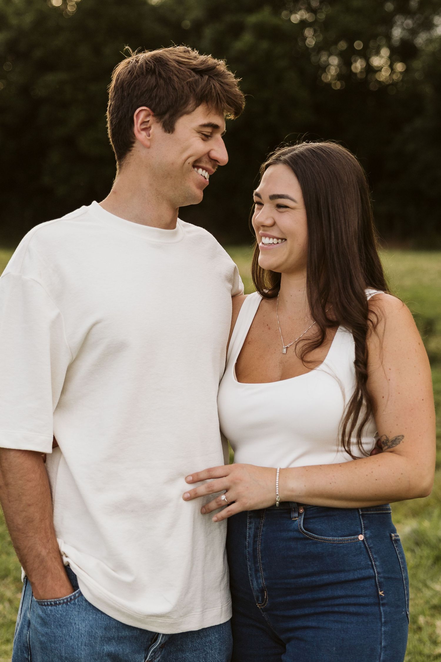 Smiling couple standing together in a green field, both wearing white tops and blue jeans, looking at each other happily.
