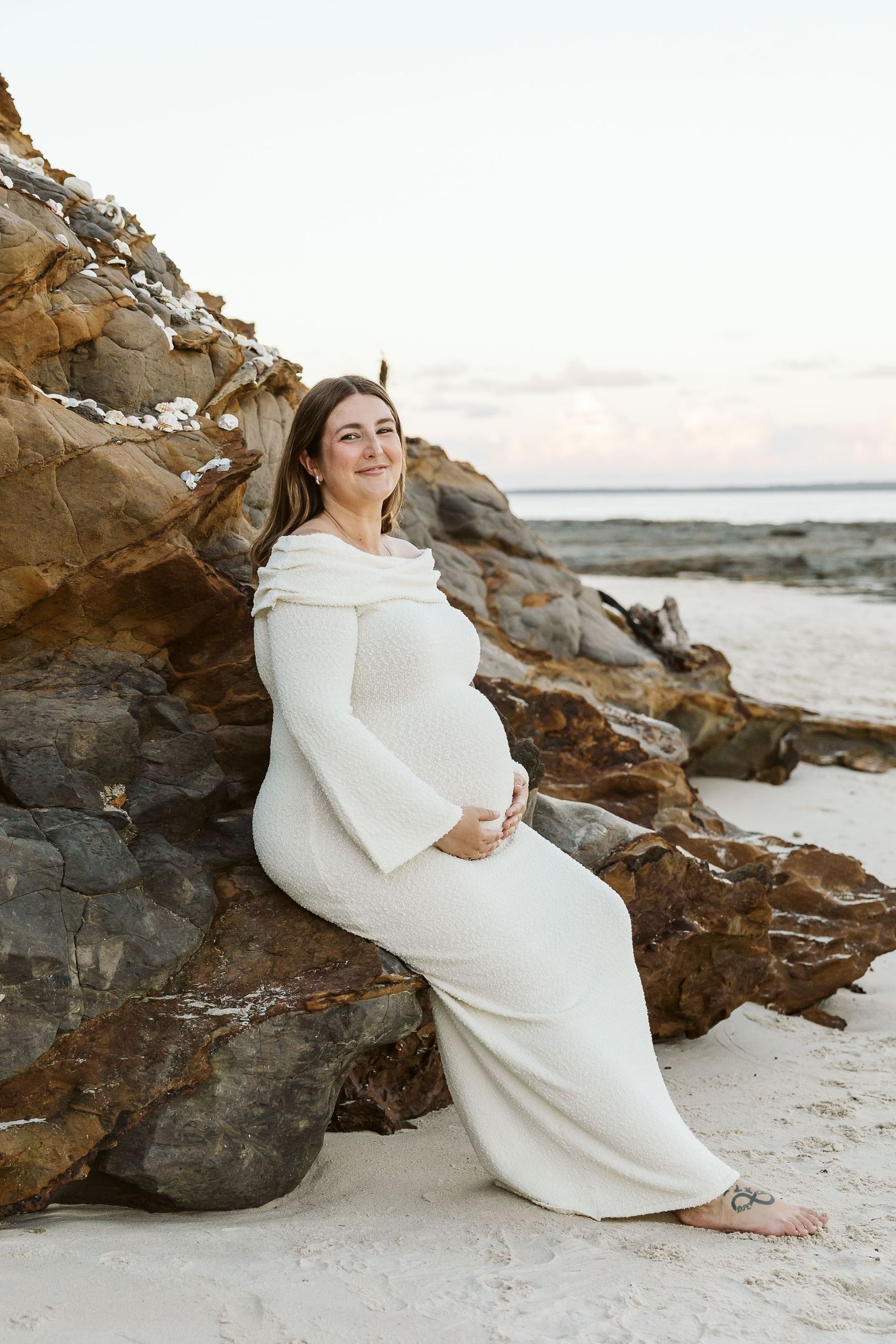 Radiant pregnant woman in white bell-sleeve dress sitting against coastal rocks on a sandy beach at sunset.
