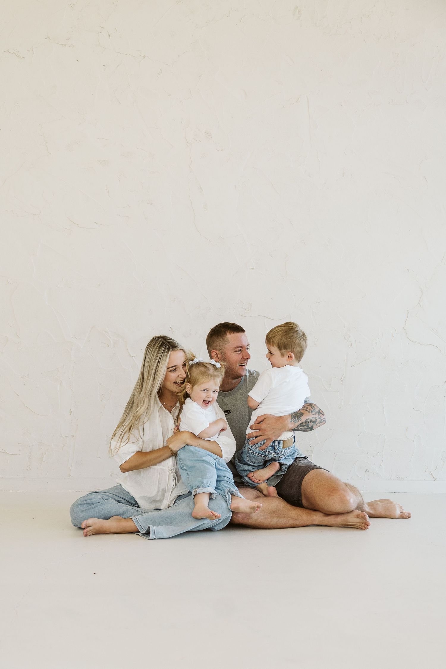 Happy family of four sitting on studio floor, parents laughing with two young children in casual clothing.