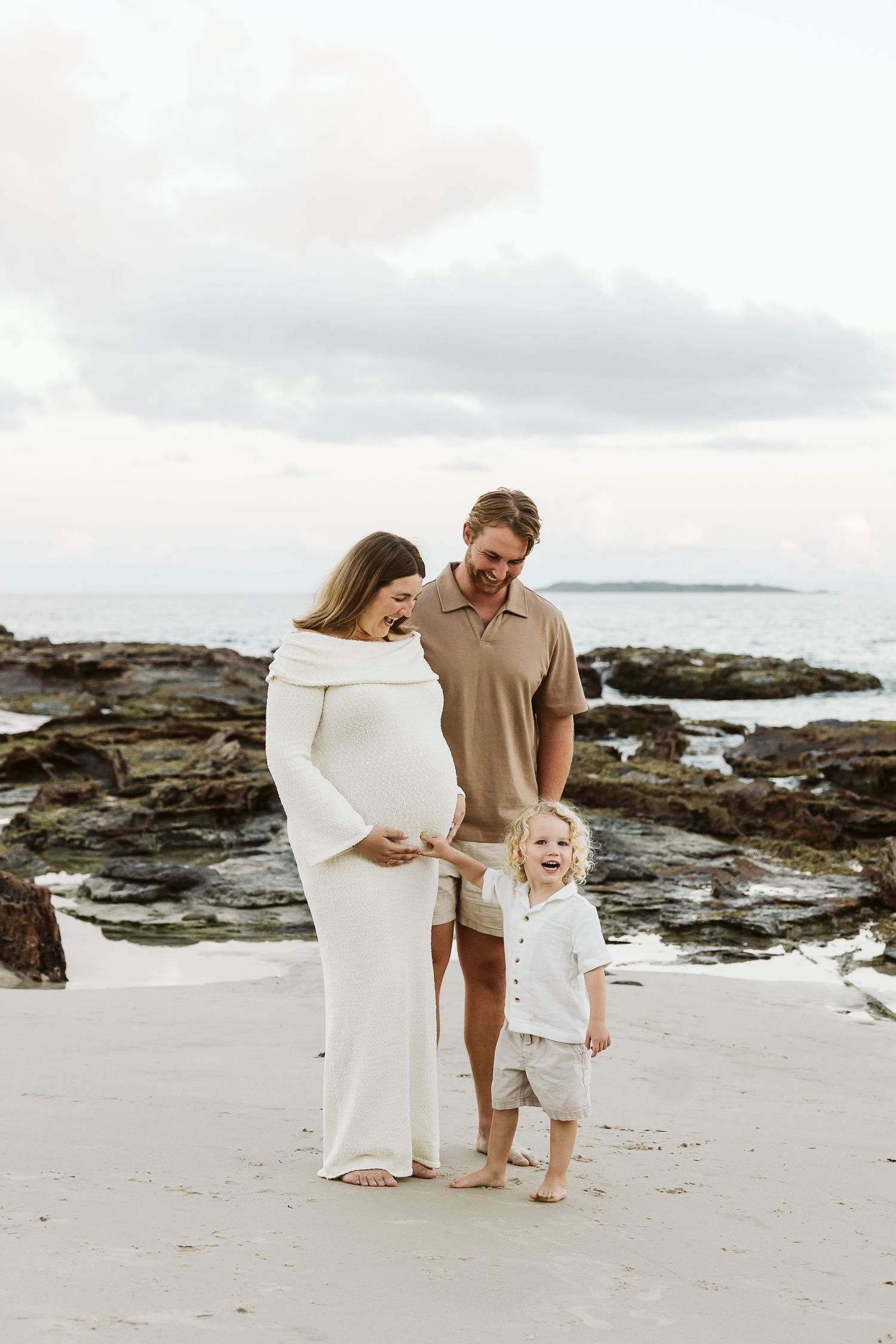 Pregnant couple with toddler son posing on rocky beach at sunset.