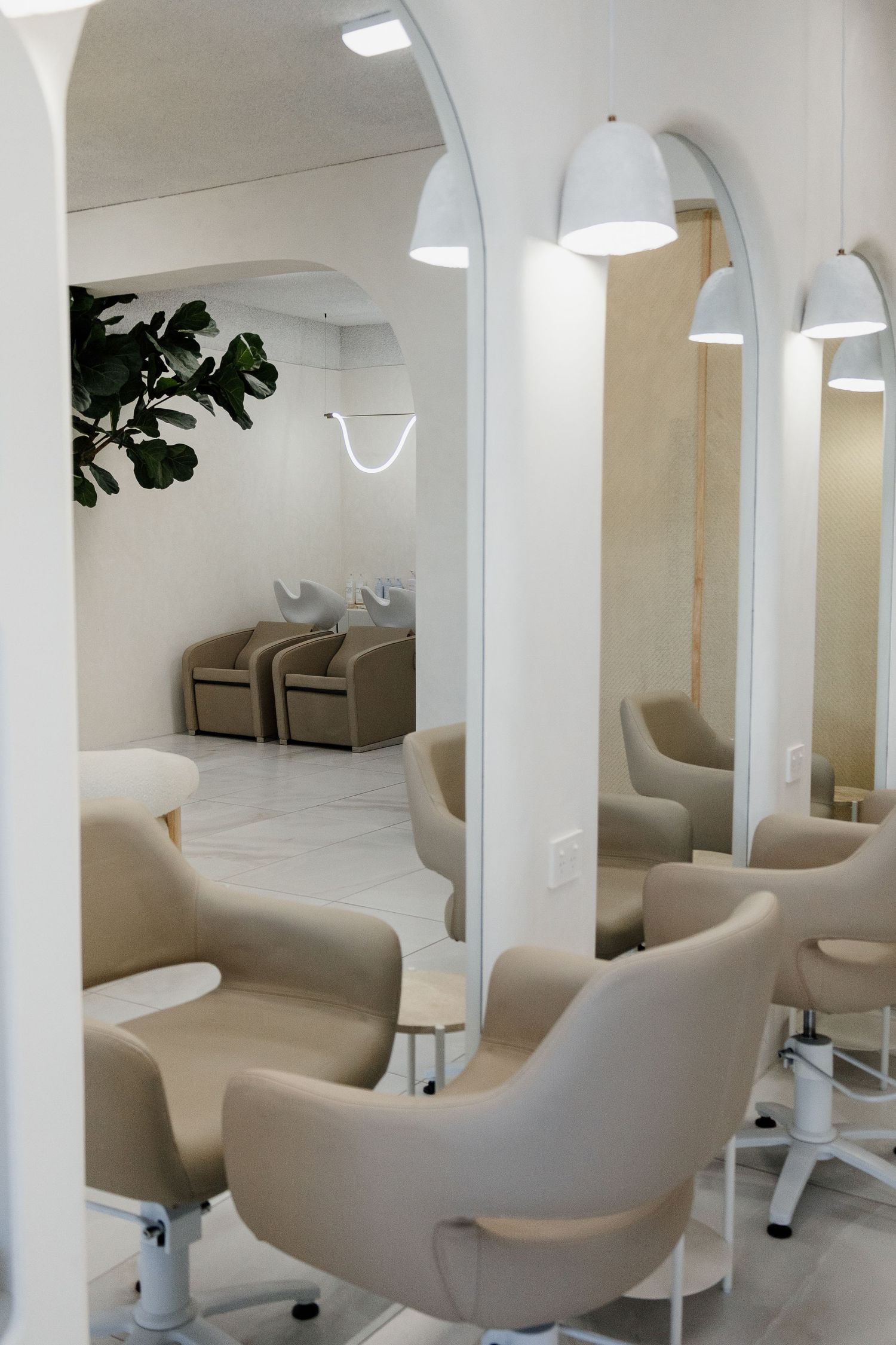 Bright modern hair salon interior with beige styling chairs, arched mirrors, and pendant lighting.