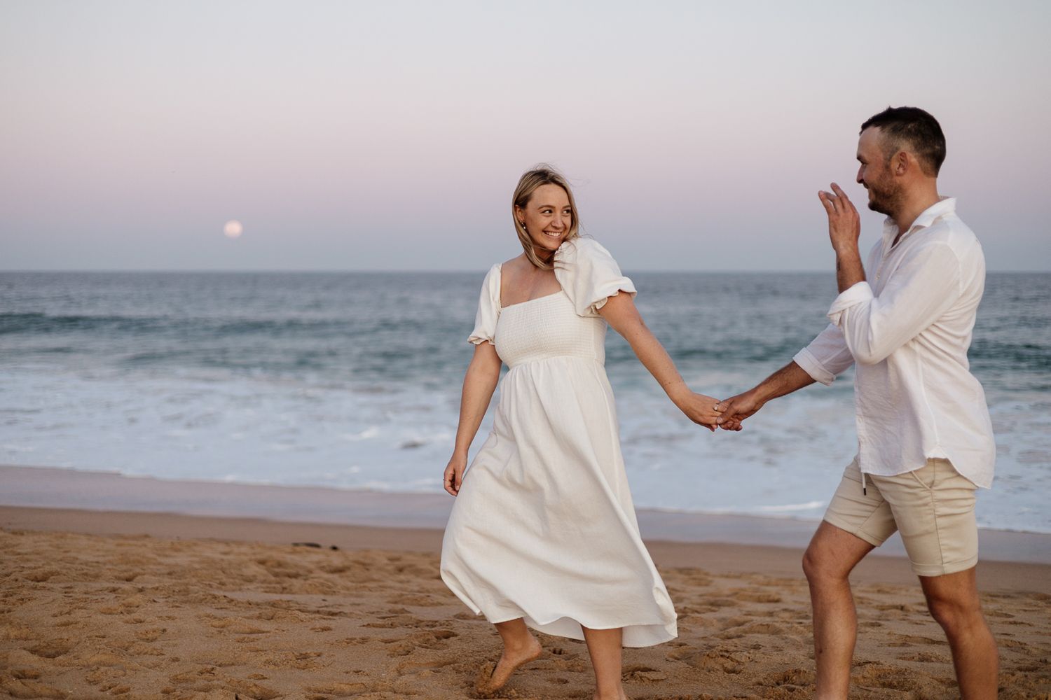 Couple in white outfits holding hands and walking on a sandy beach at sunset with ocean waves.