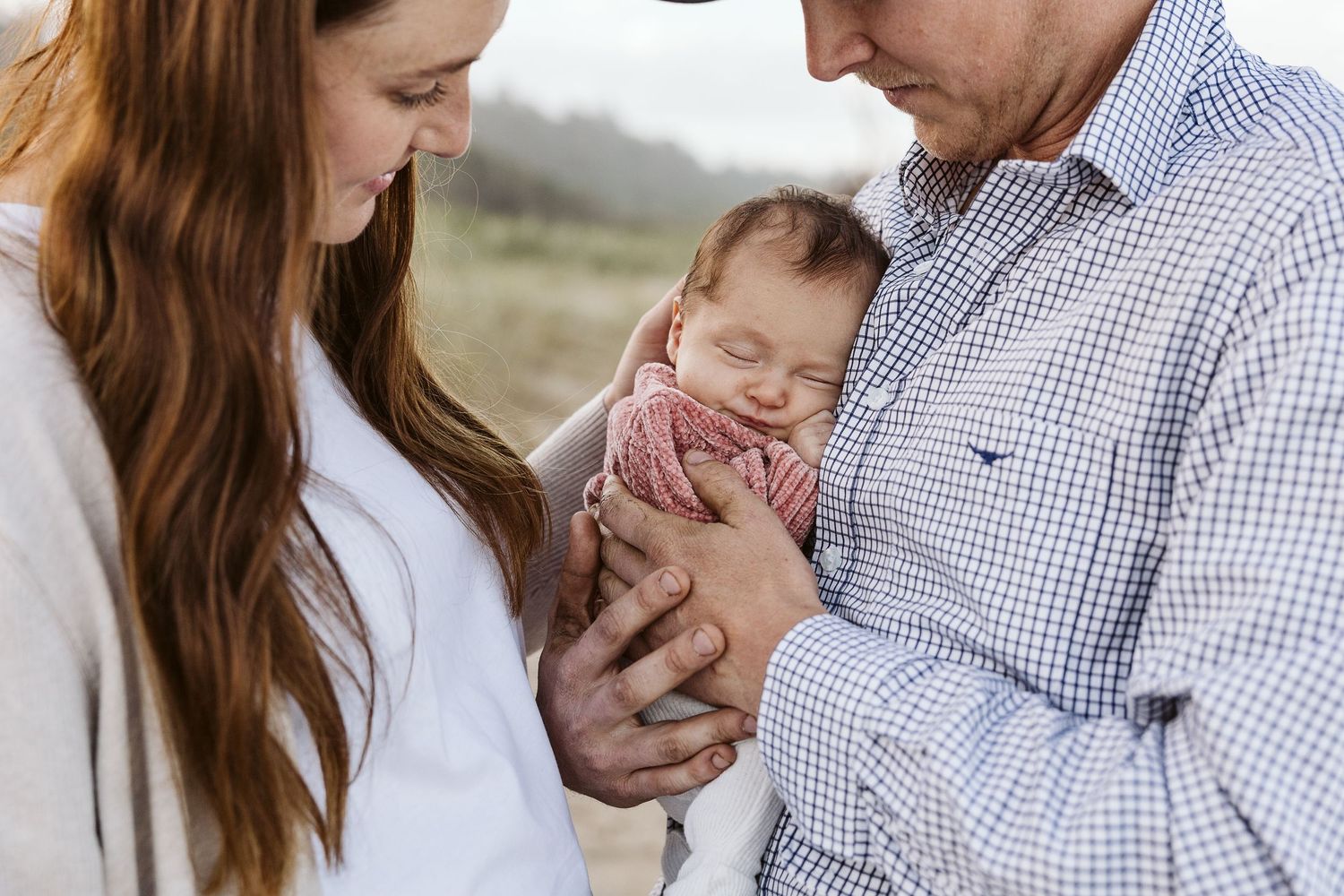 New parents tenderly holding their sleeping newborn baby wrapped in a pink blanket outdoors.