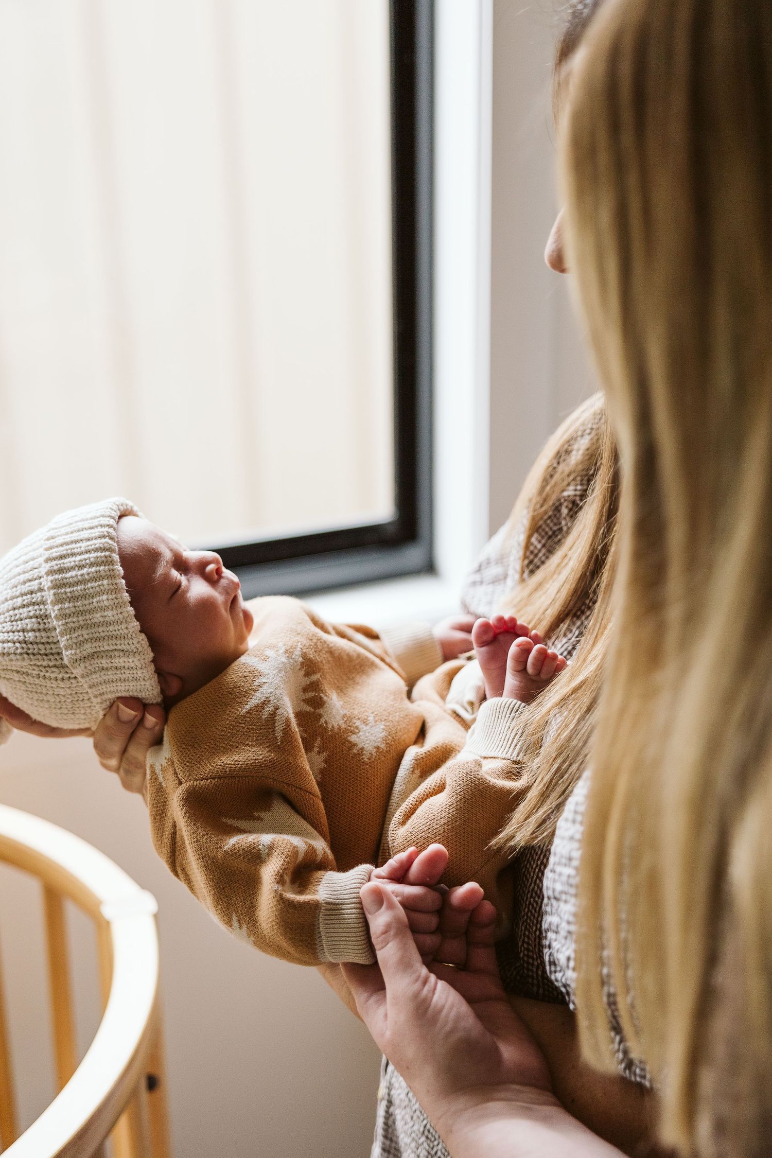 Blonde mother holding sleeping newborn in tan outfit and knit hat near nursery window.