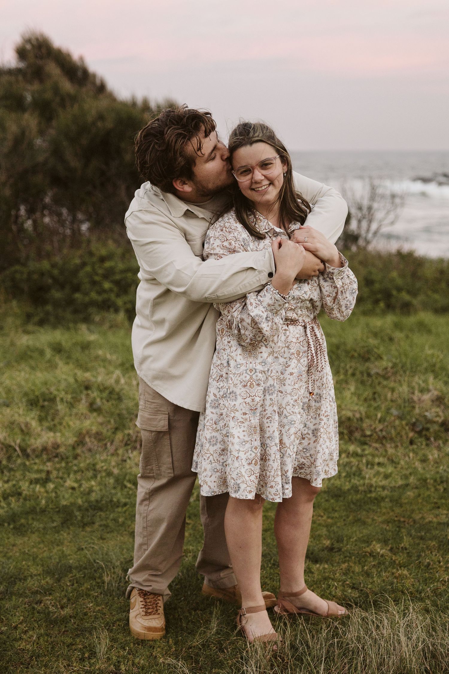 Man hugging and kissing woman in floral dress while she smiles, standing on grassy coastal cliff.