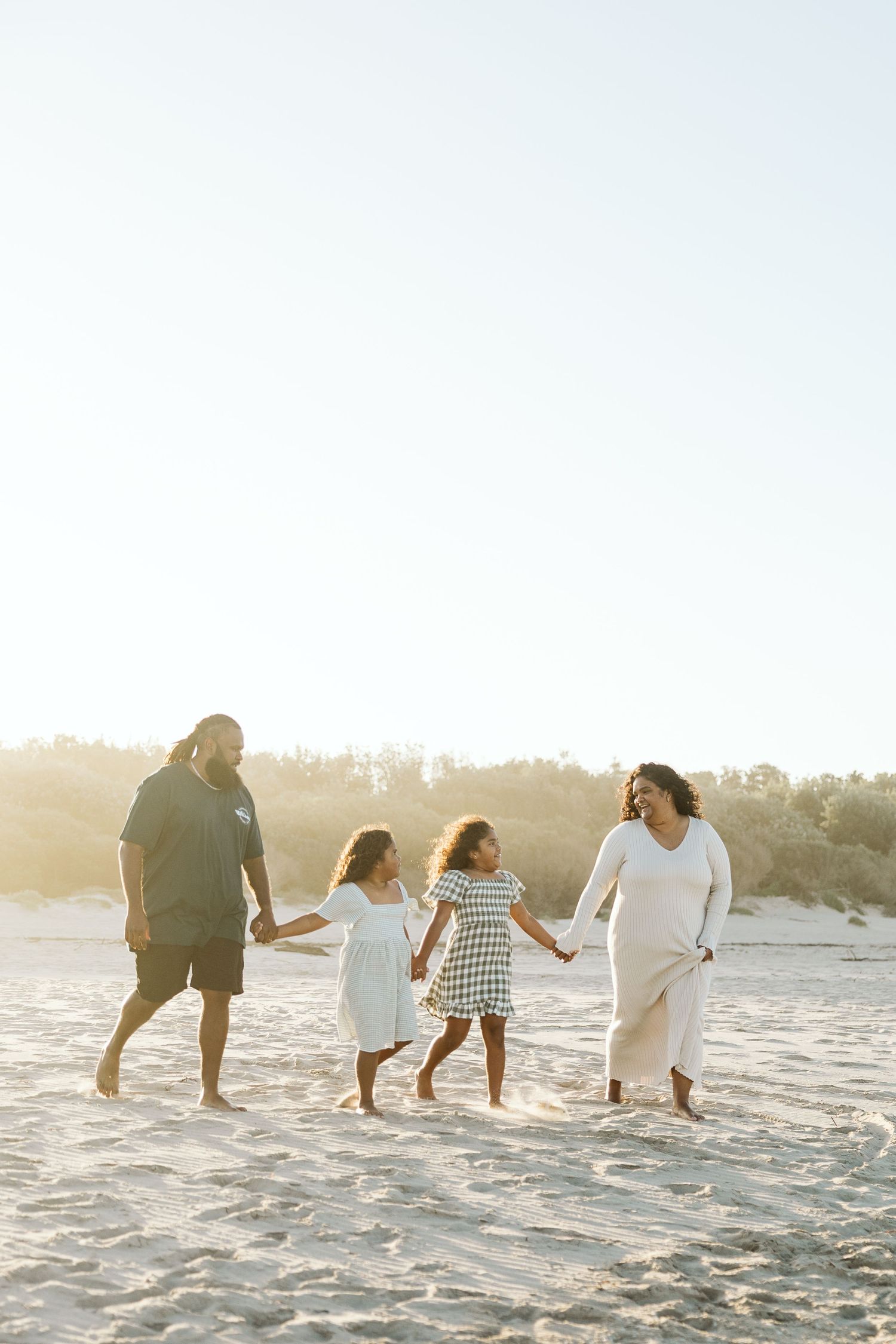 Family of four walking hand in hand on a sunny beach, parents and two young girls in white dresses.