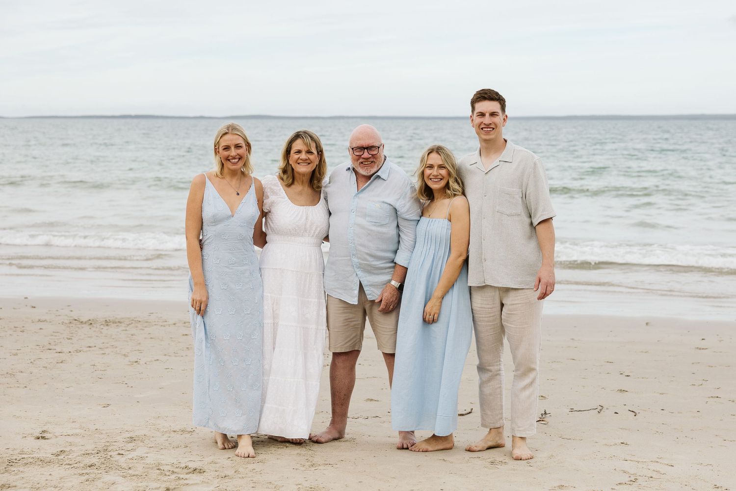 Family of five smiling together on a sandy beach, wearing coordinated light blue and neutral outfits with ocean in background.