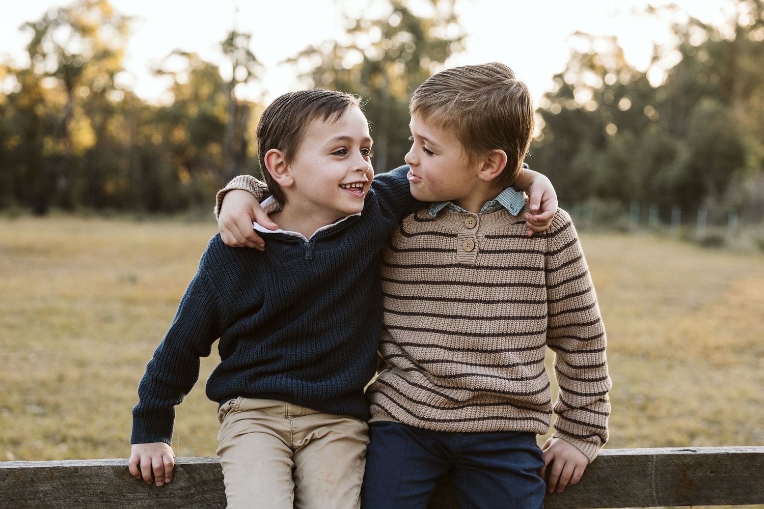 Two young boys sitting on a fence smiling at each other outdoors in a field.