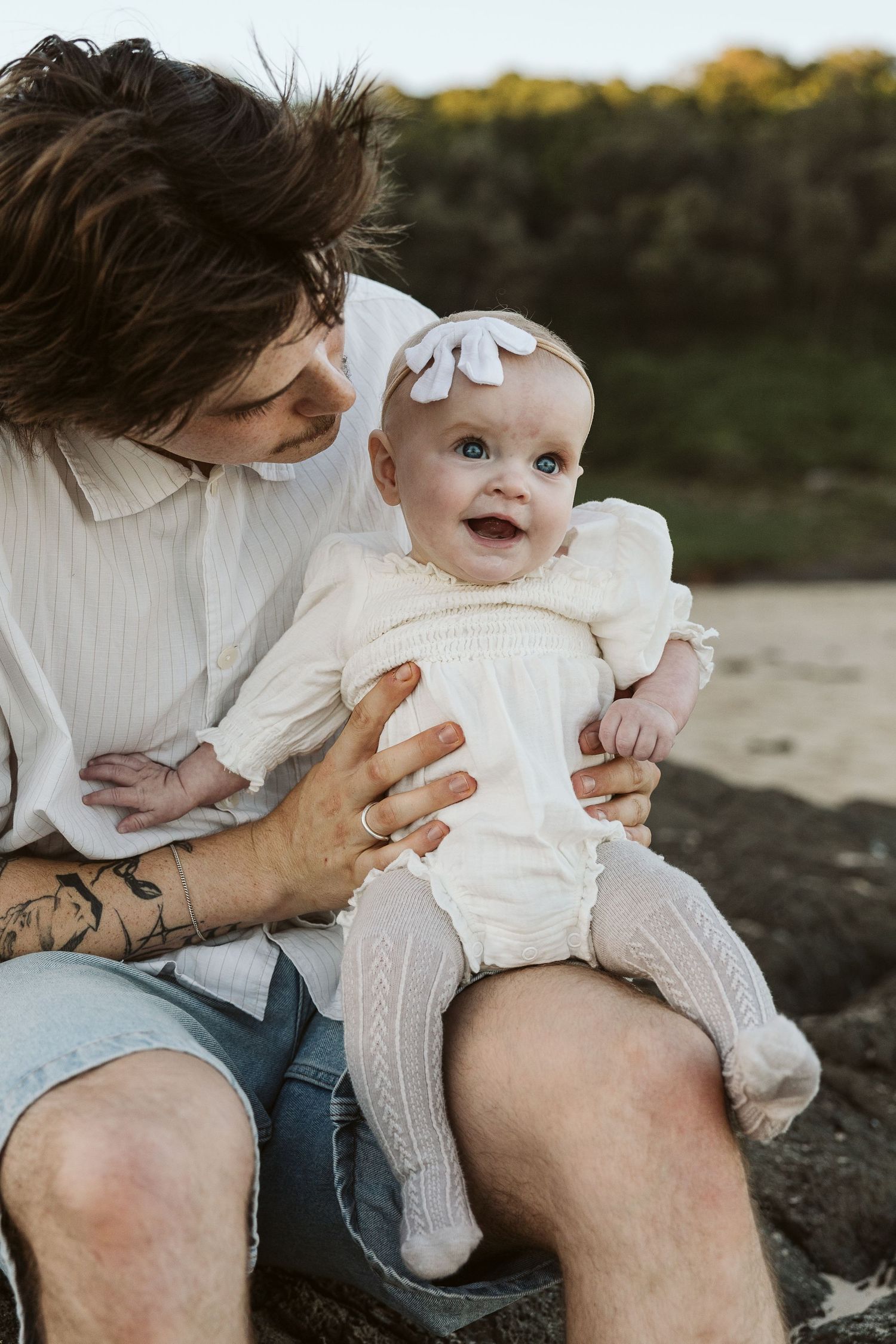 A tattooed father sits outdoors holding his smiling baby girl in a white romper.