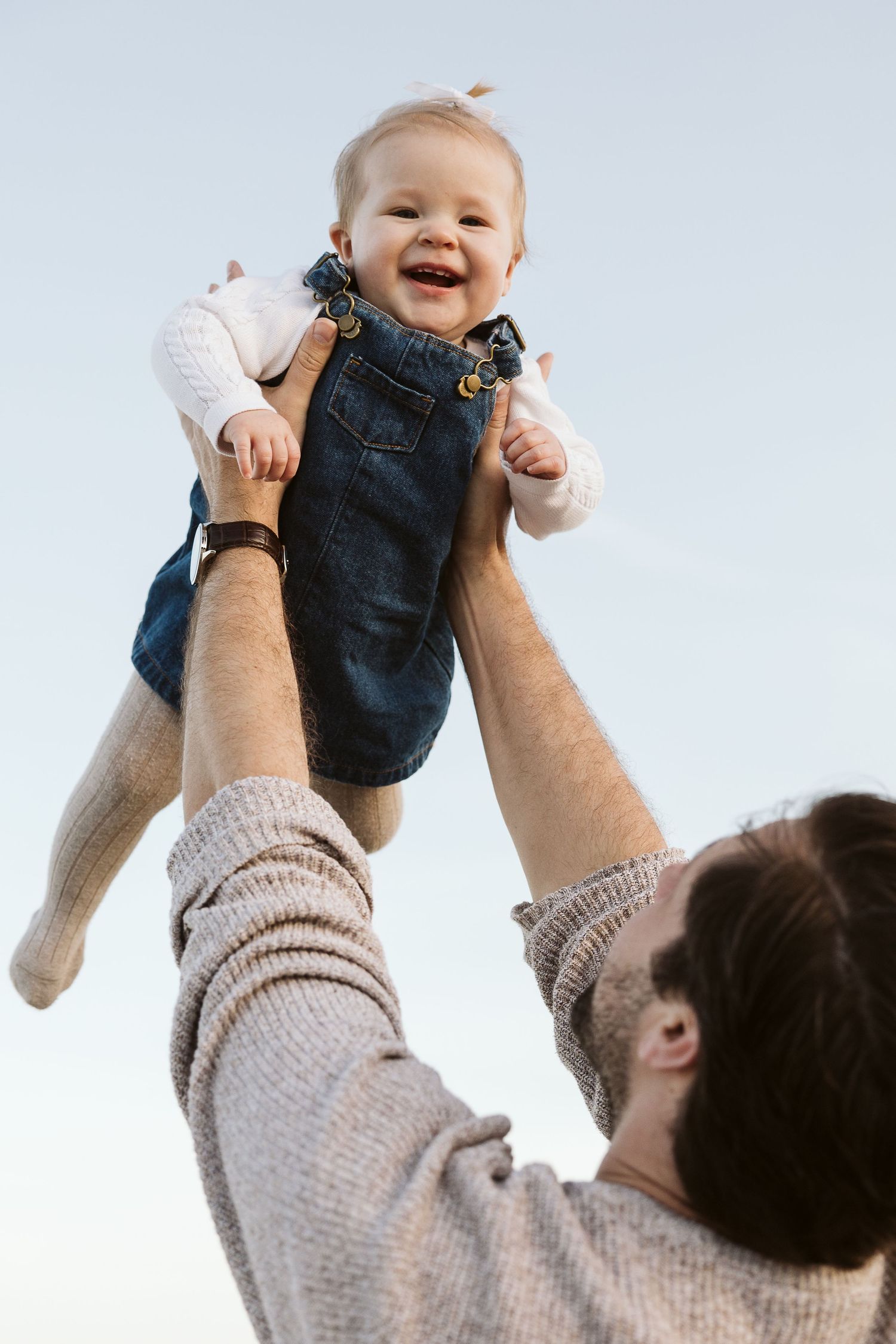 Happy baby in denim overalls being lifted high in the air by father against a clear sky.