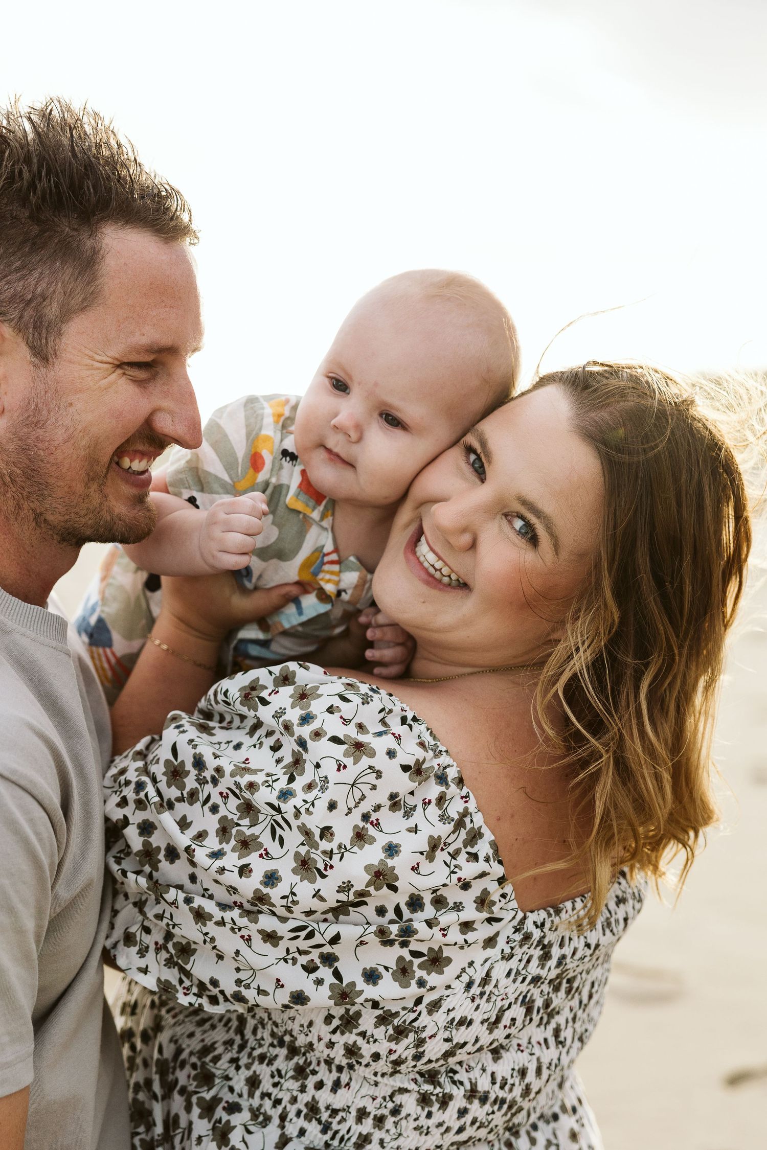 Happy family with smiling parents holding their baby at the beach in warm sunlight.