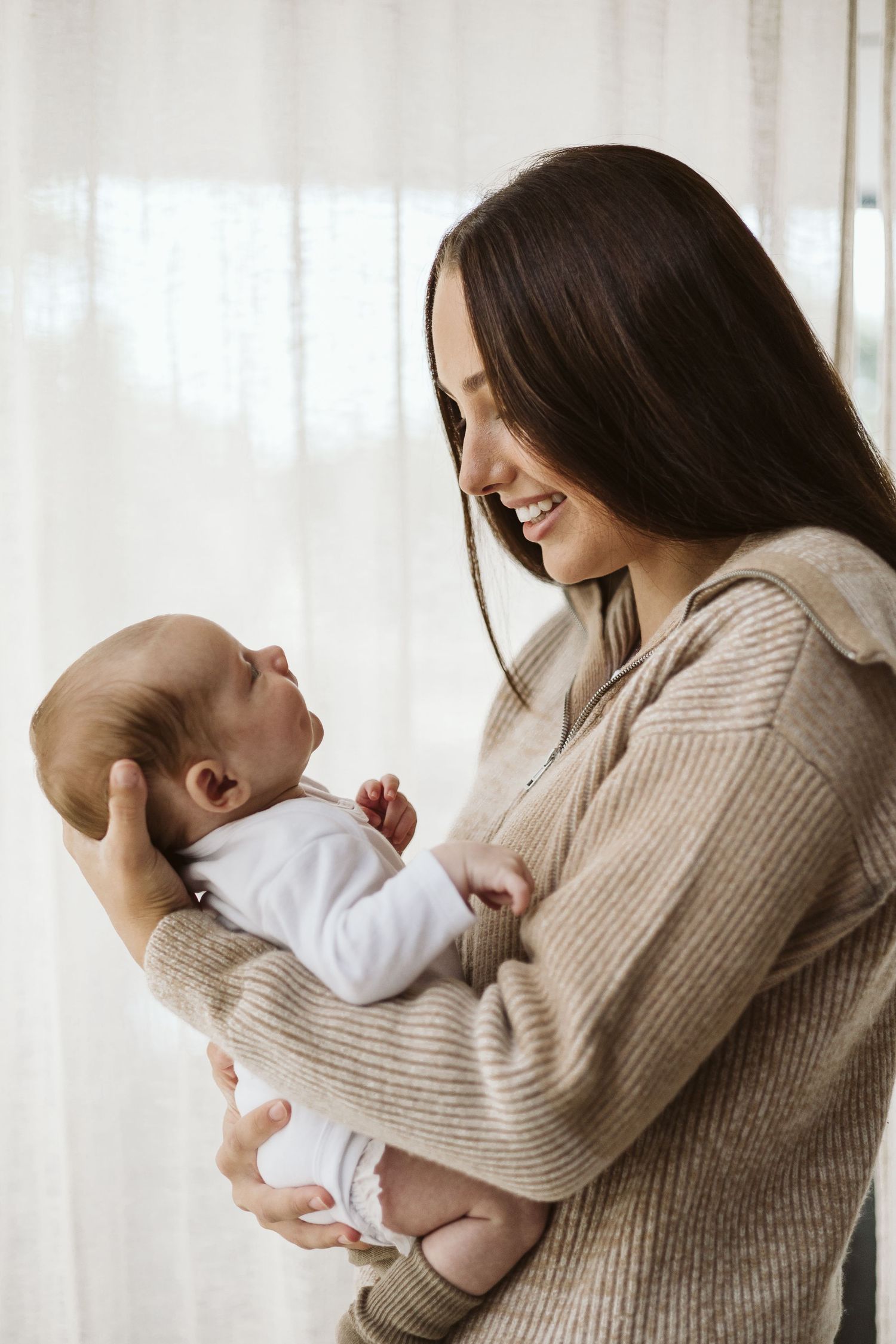 Smiling dark-haired mother in beige sweater holds her newborn baby lovingly.