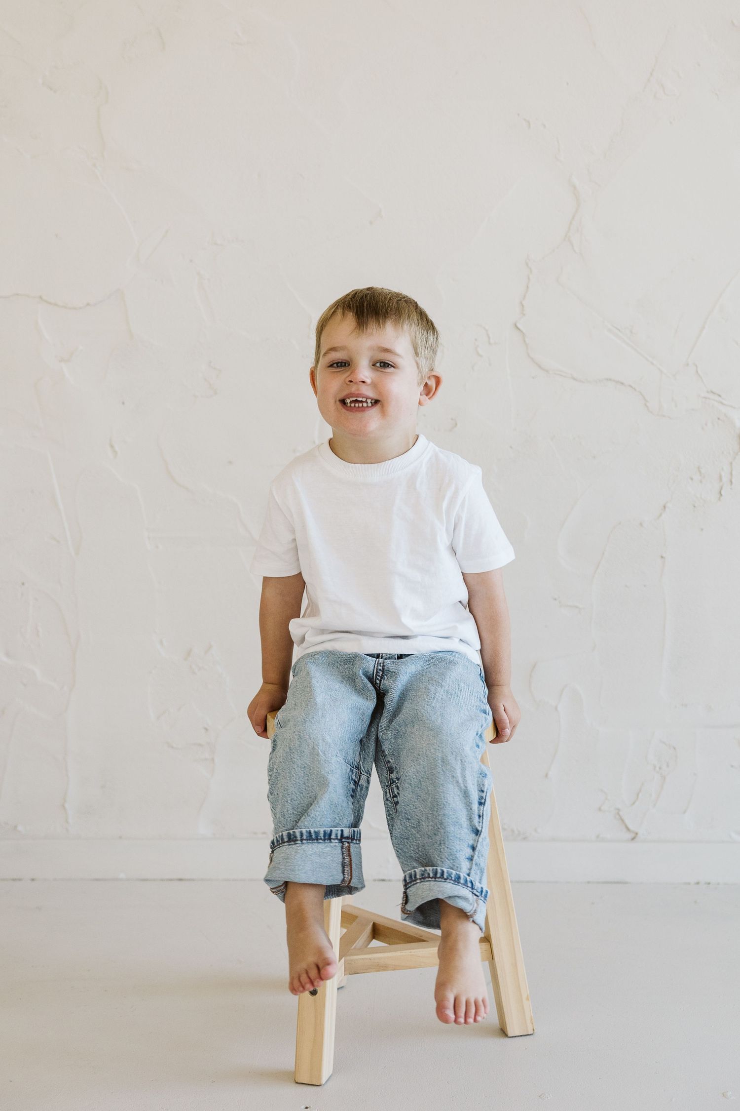 A cheerful young boy in a white t-shirt and jeans sits barefoot on a wooden stool against a white wall.