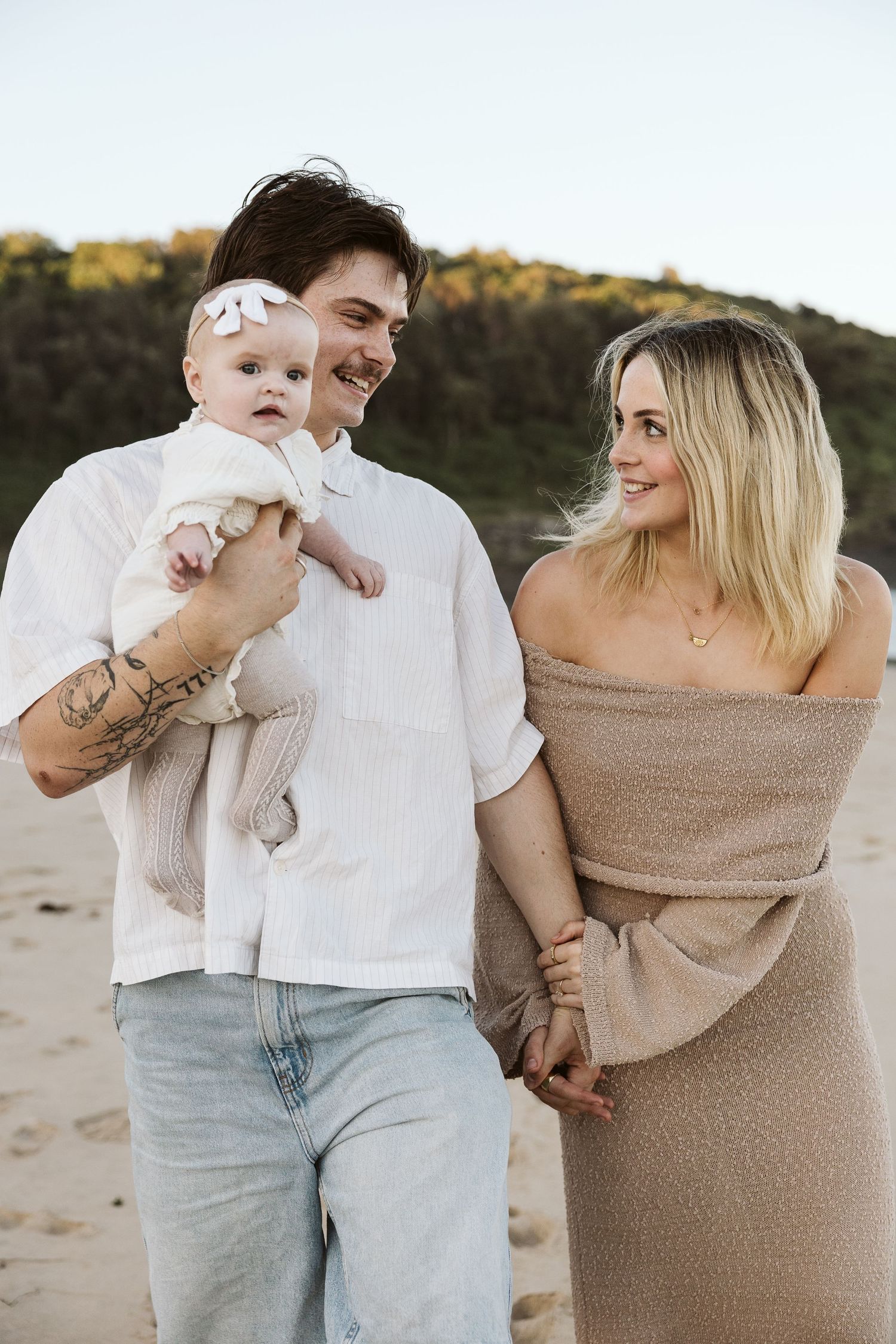 Smiling tattooed father holding baby girl in white dress with blonde mother on sandy beach.
