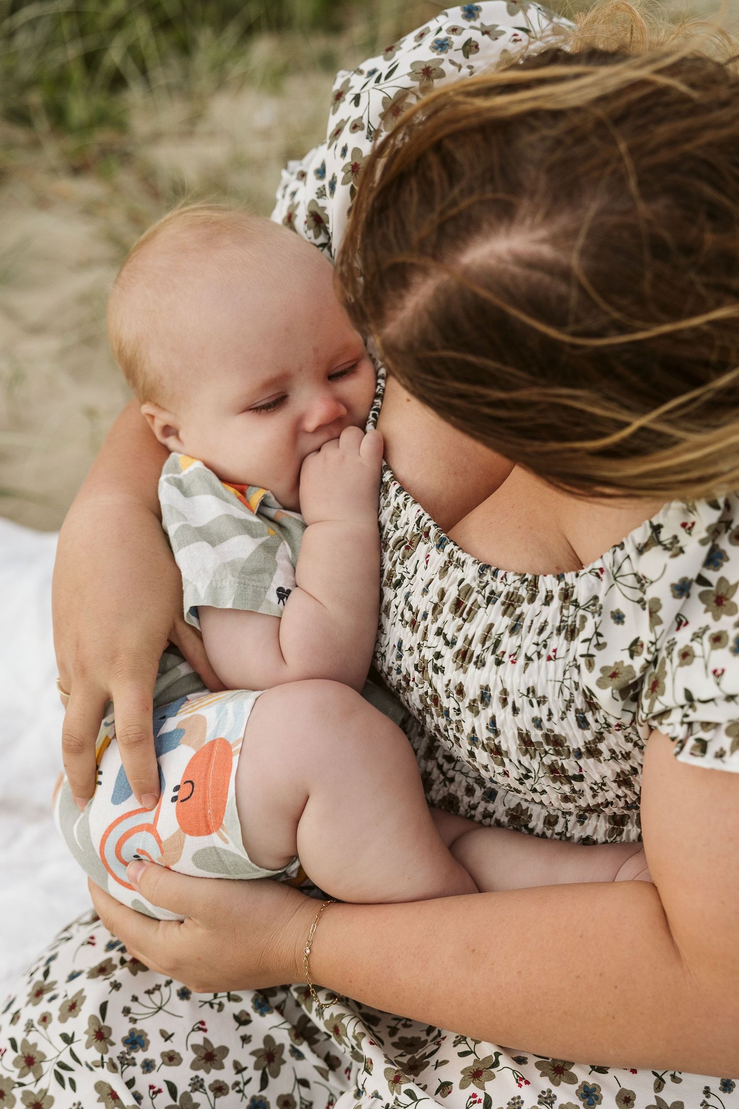 Mother in floral dress tenderly holding and cuddling her sleepy infant baby outdoors in natural setting.