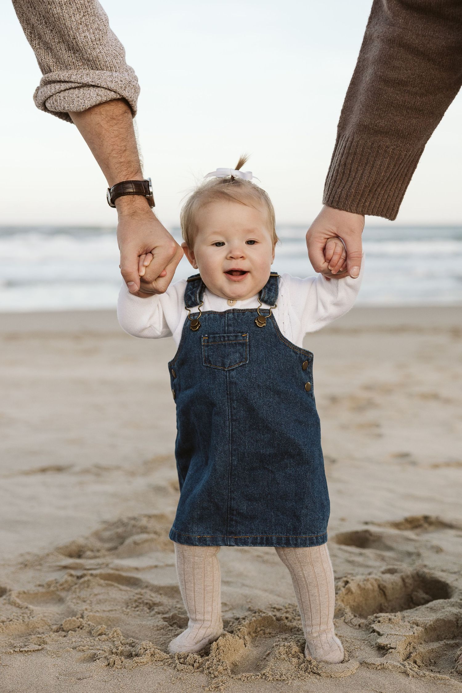 Smiling baby girl in denim dress taking first steps on sandy beach while being supported by two adults holding her hands.