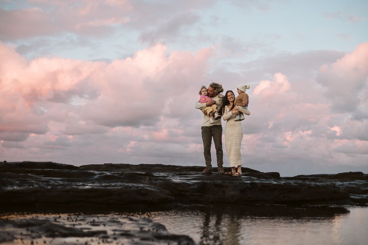 Family of four standing on rocks at sunset with pink sky. Winter photoshoot, coastal location.