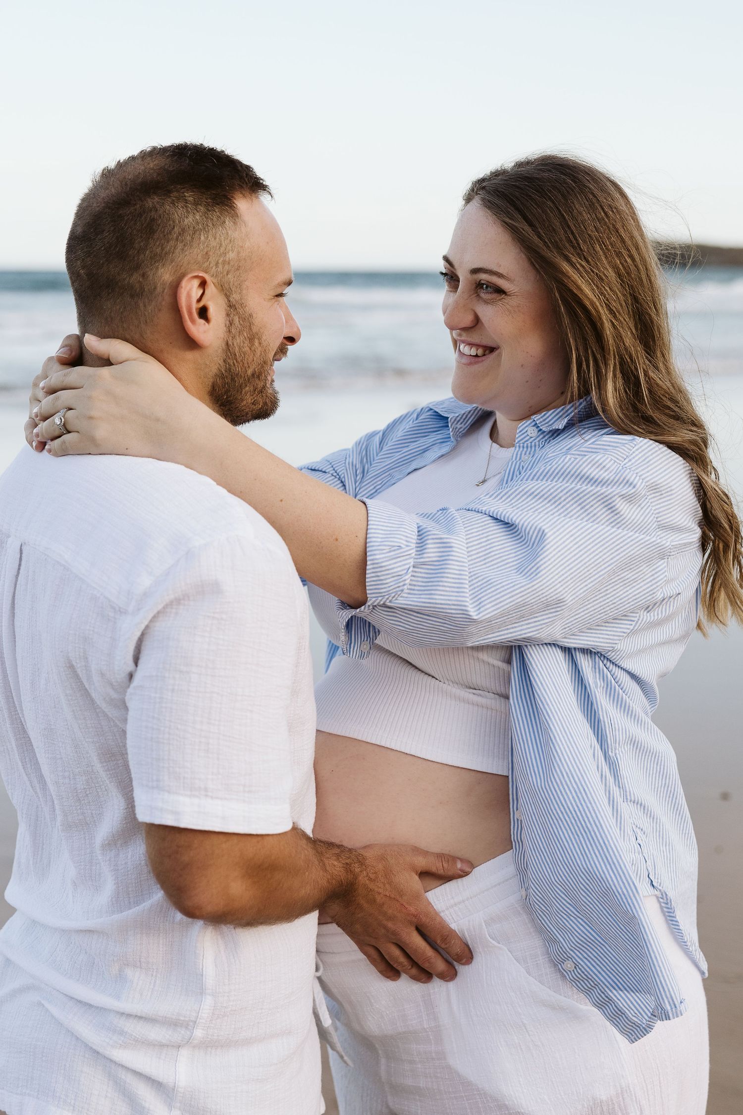 Smiling pregnant woman in blue shirt embracing bearded man in white on sandy beach at sunset.