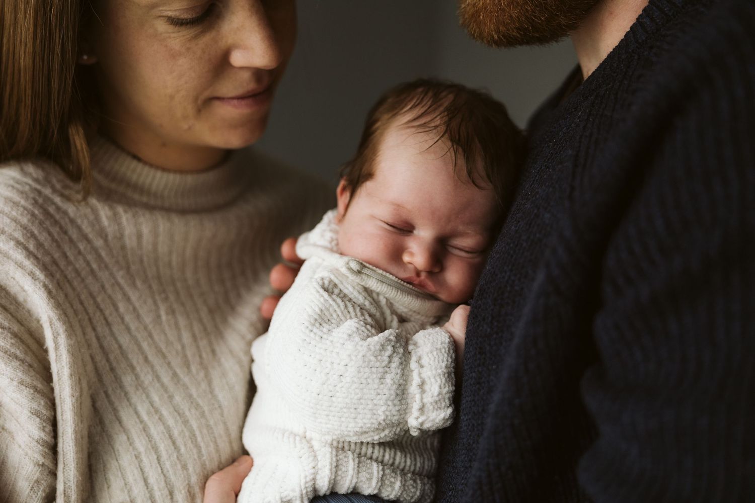 Parents in cozy sweaters lovingly hold their sleeping newborn baby together.