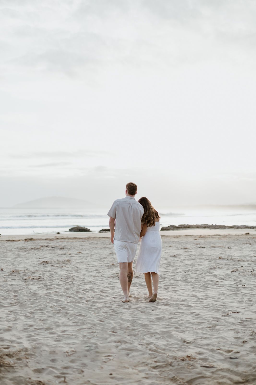 Couple dressed in white walking hand in hand along a sandy beach under a cloudy sky.