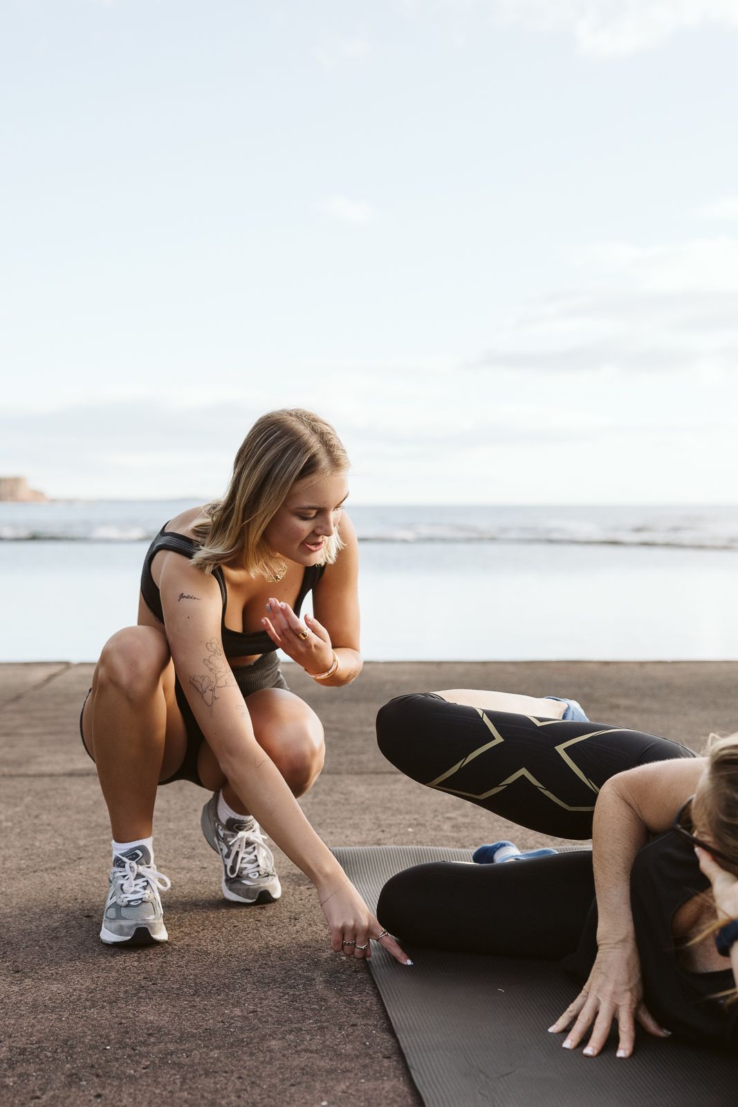 Blonde fitness trainer crouches to assist client with exercise on outdoor oceanside mat.