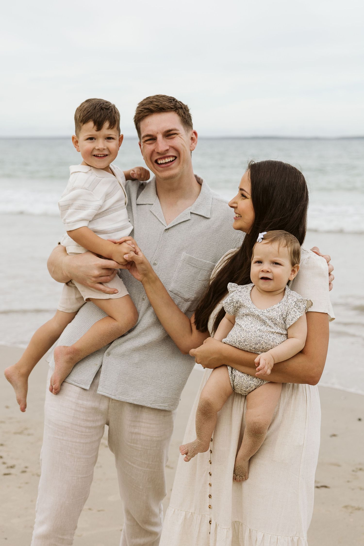 Happy young family of four posing on a sandy beach, parents holding two toddlers, smiling in neutral tones.