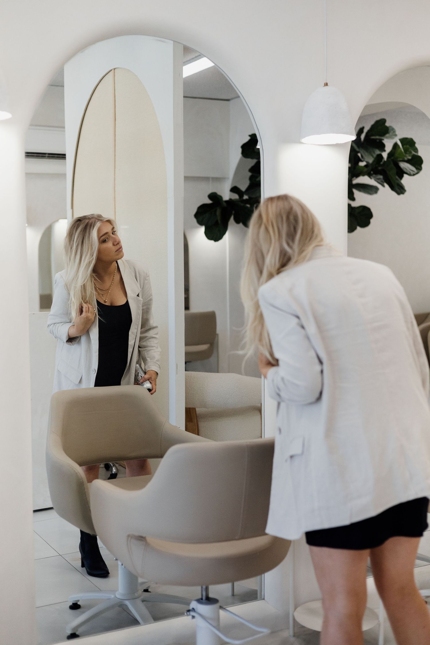 Blonde woman in white blazer examining her hair in large arched mirror at modern hair salon.