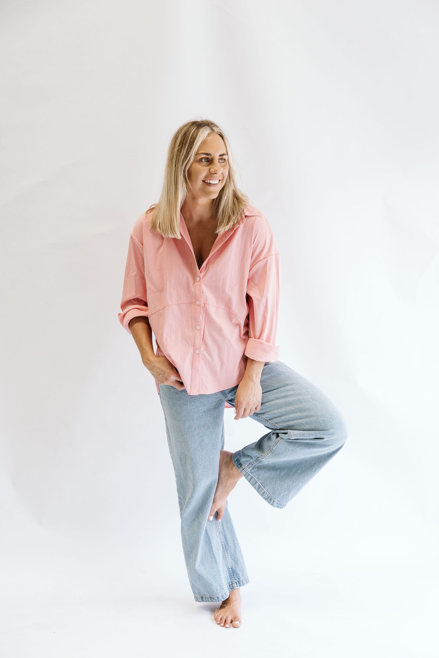 Smiling blonde woman in pink shirt and wide-leg jeans poses on white background.