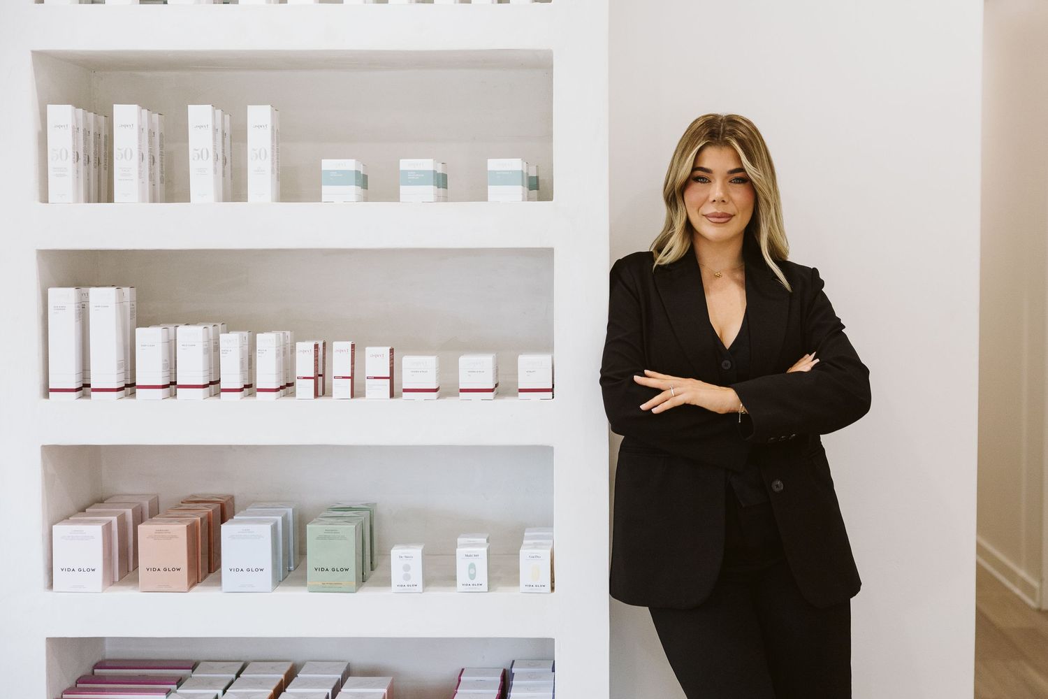 Confident woman in black blazer standing beside white shelves stocked with skincare products at an aesthetics clinic.