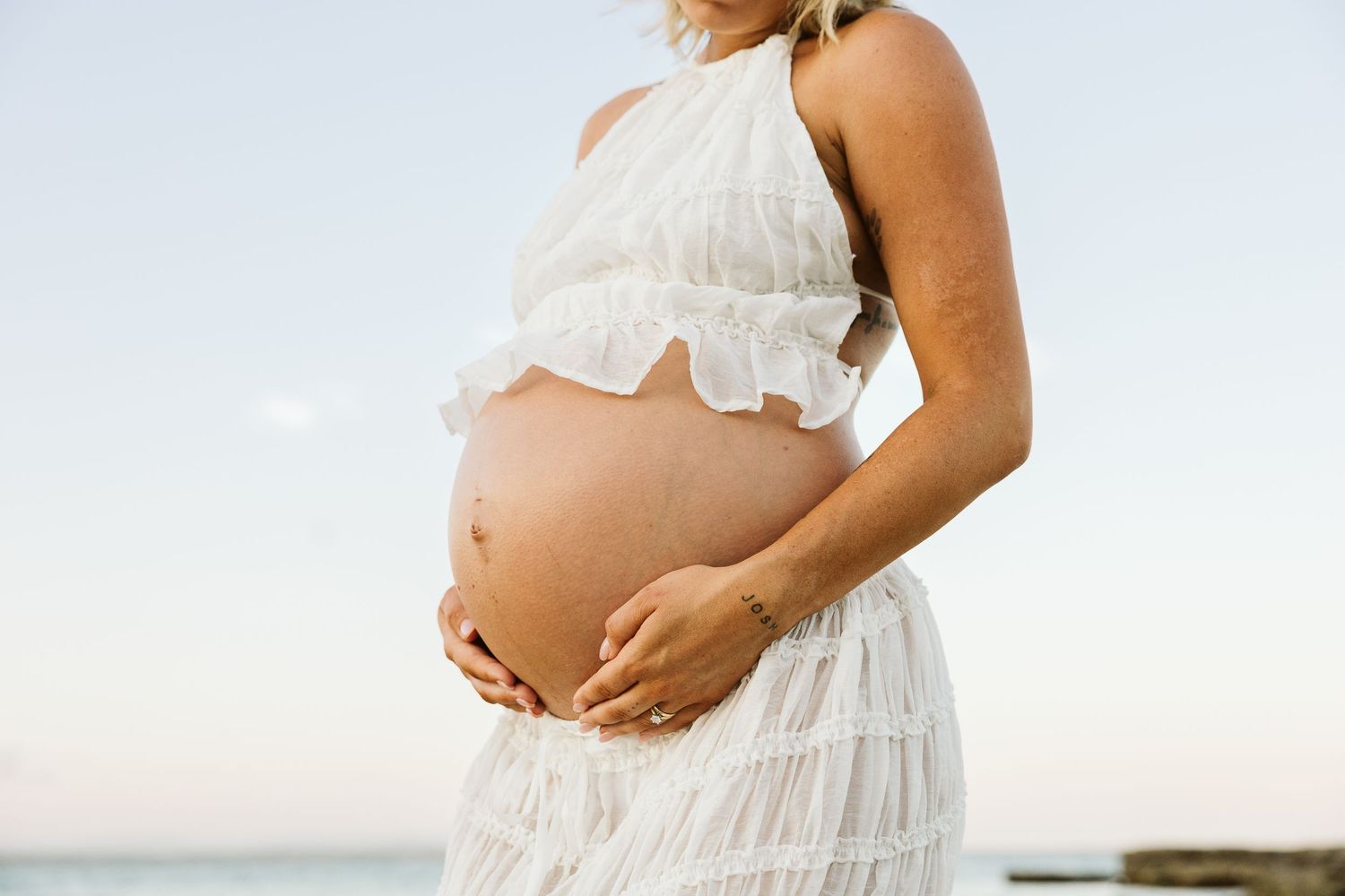 Close-up of pregnant woman in white ruffled crop top and skirt gently holding her bare baby bump at the beach.
