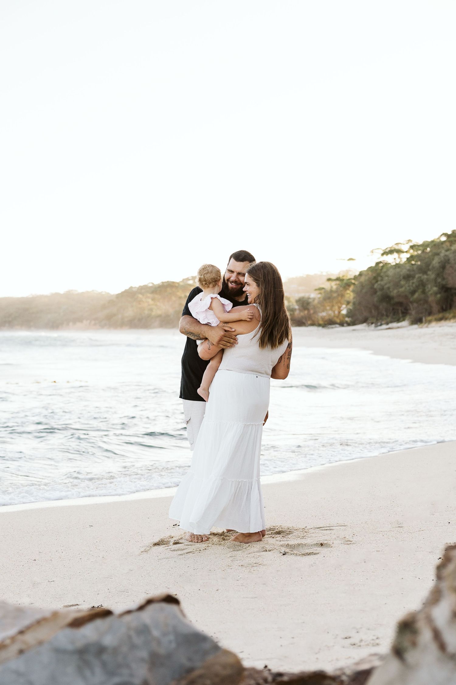 Young couple holding baby together on a sunlit beach at golden hour.