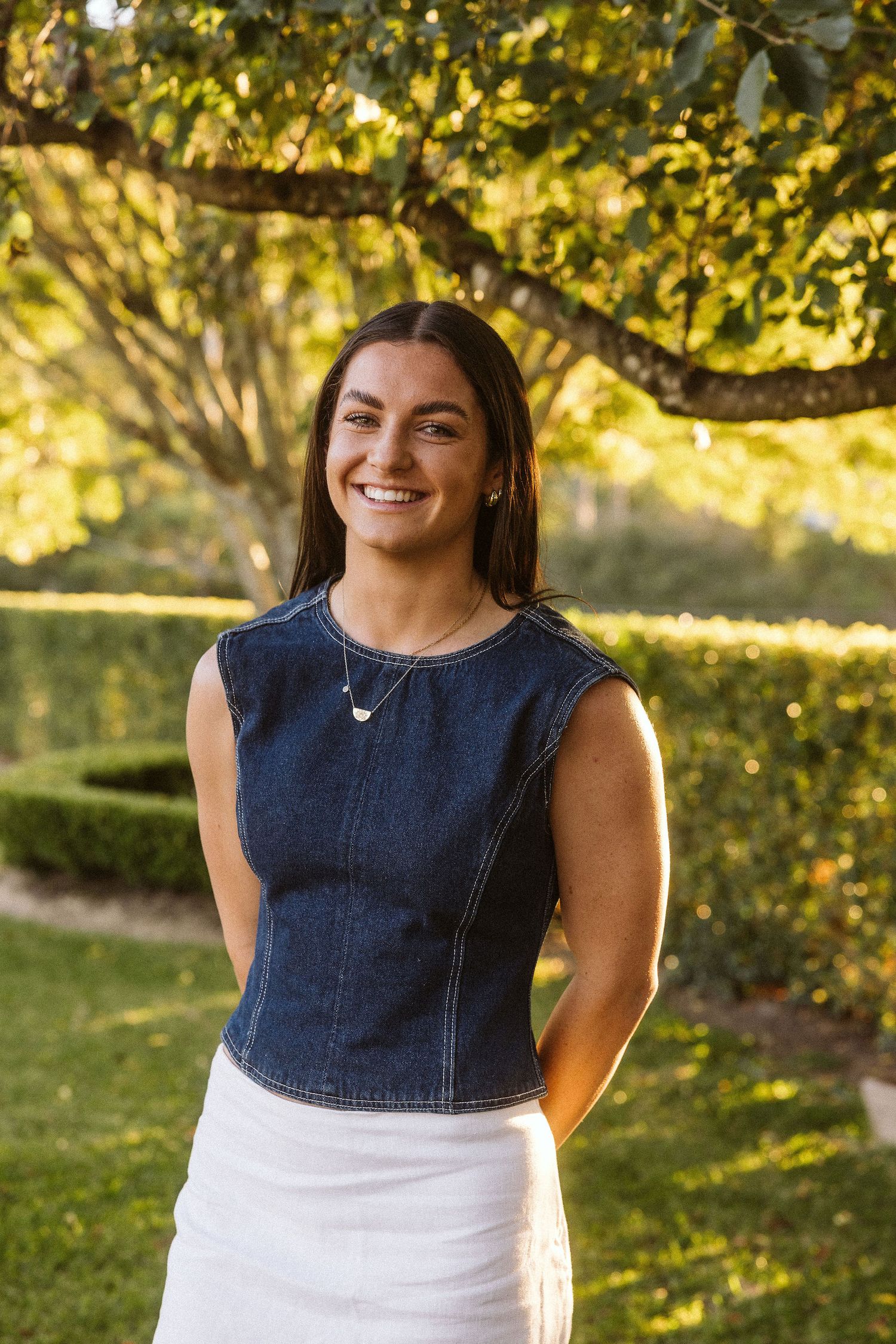 Smiling brunette woman wearing denim sleeveless top and white skirt posing outdoors.