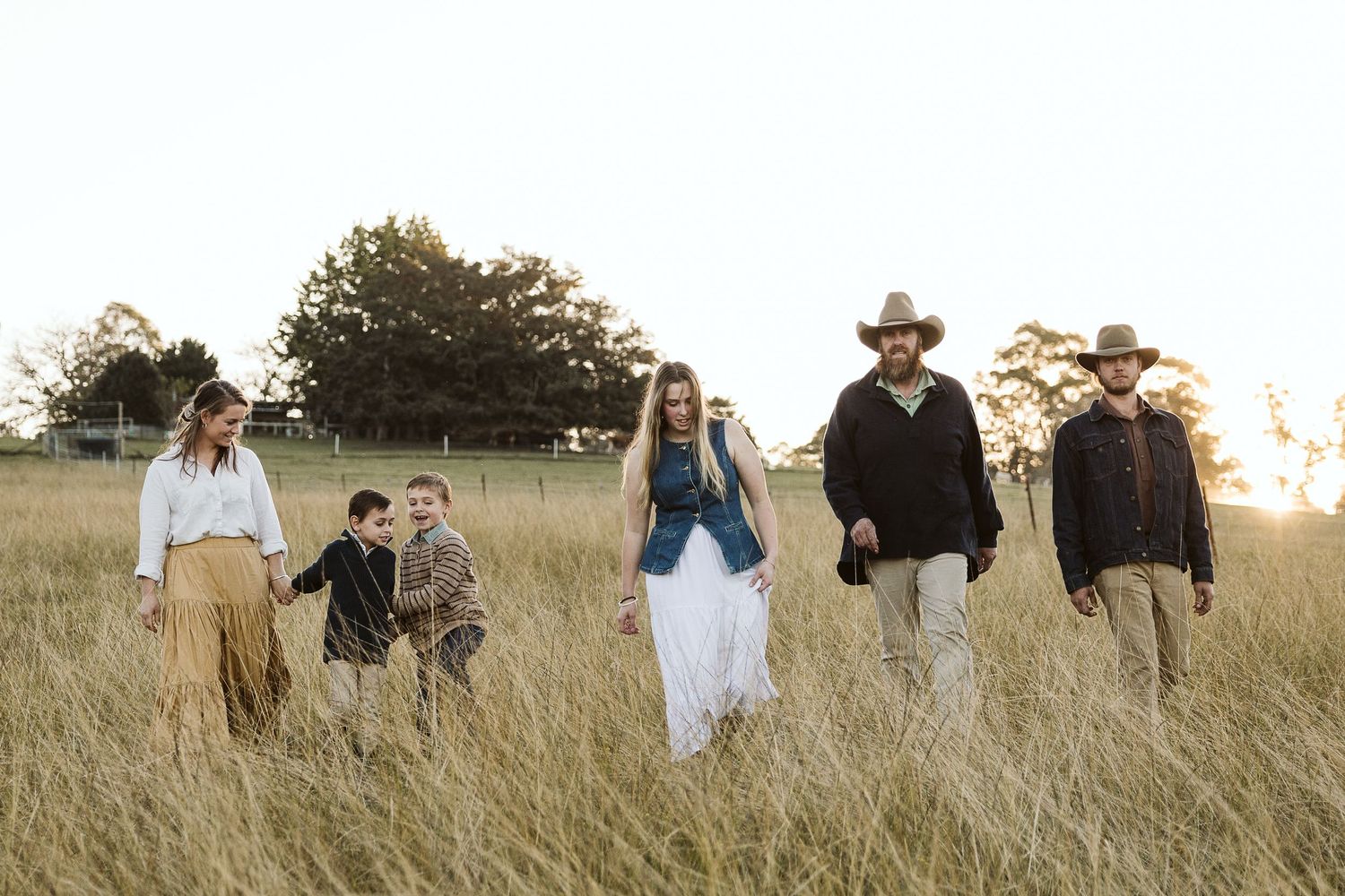A family walks through golden tall grass fields at sunset, wearing casual country attire including cowboy hats.