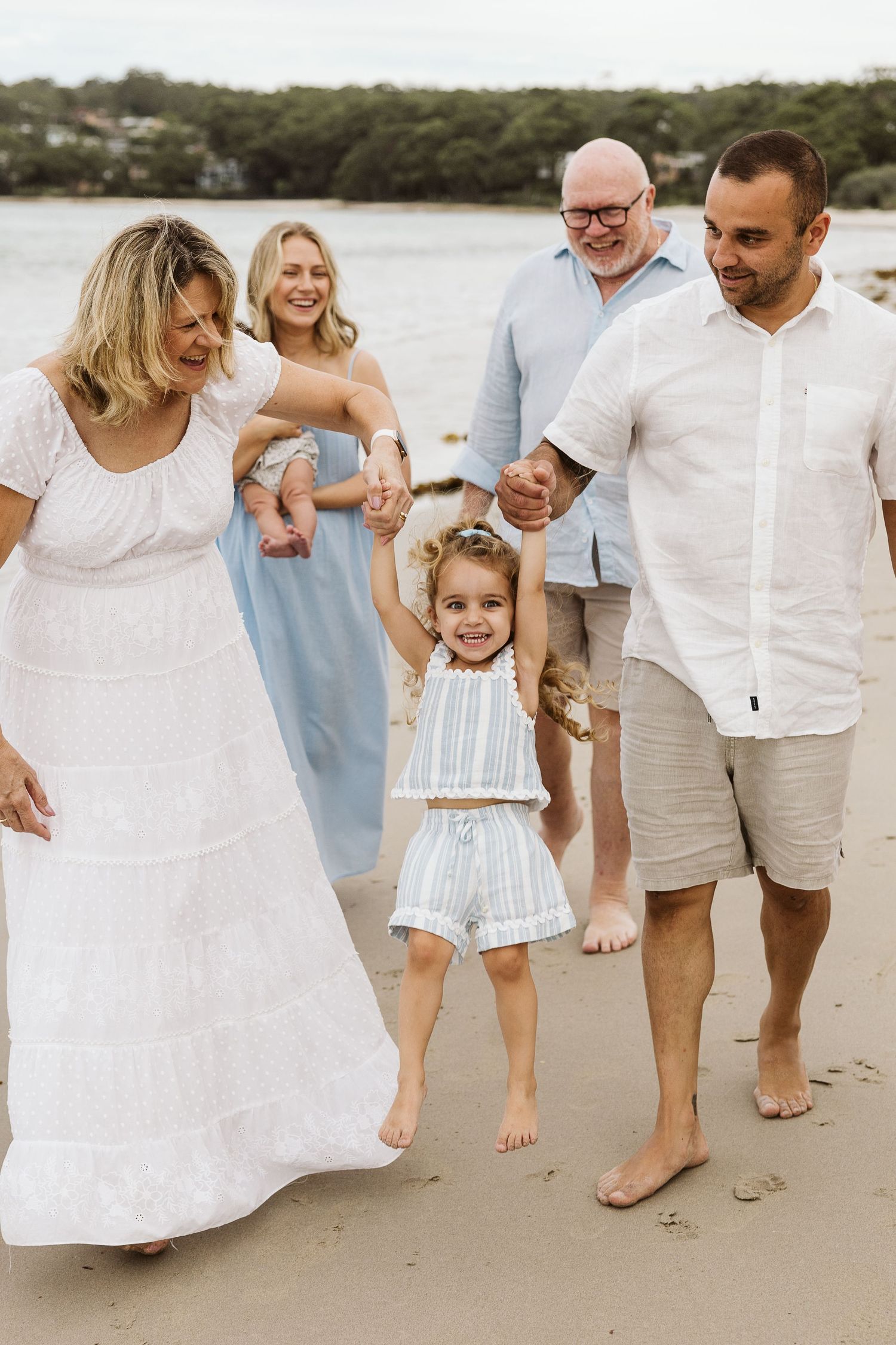 Multi-generational family walking barefoot on sandy beach, smiling and holding hands with young girl in blue striped outfit.