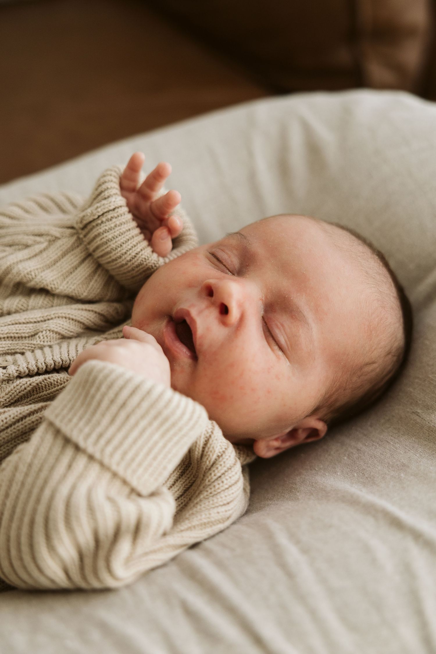 Peaceful newborn baby sleeping in a beige knit romper with hand raised in a white bassinet.