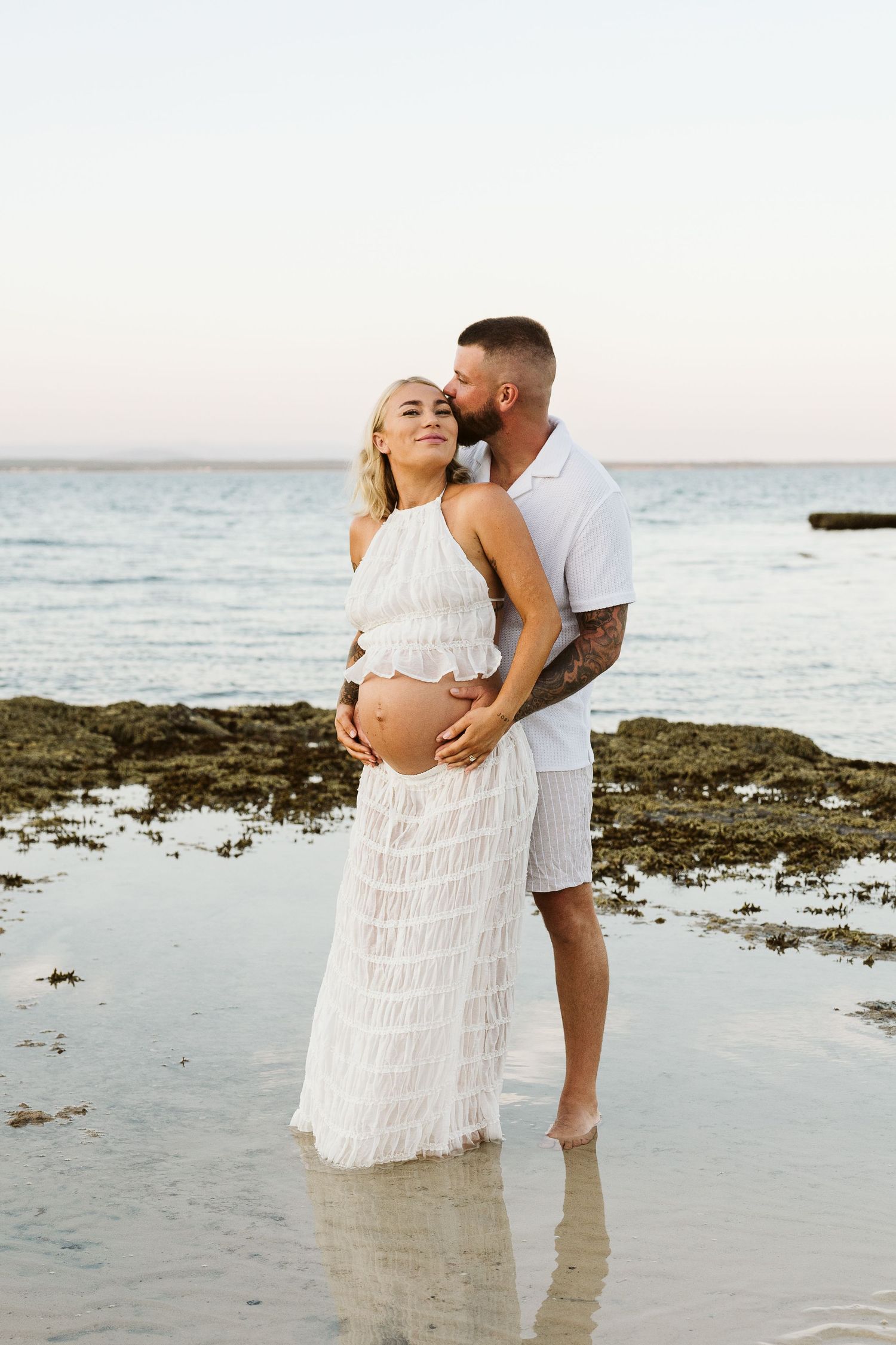 Tattooed man kissing pregnant blonde woman while cradling her bump, standing in shallow beach water at sunset.