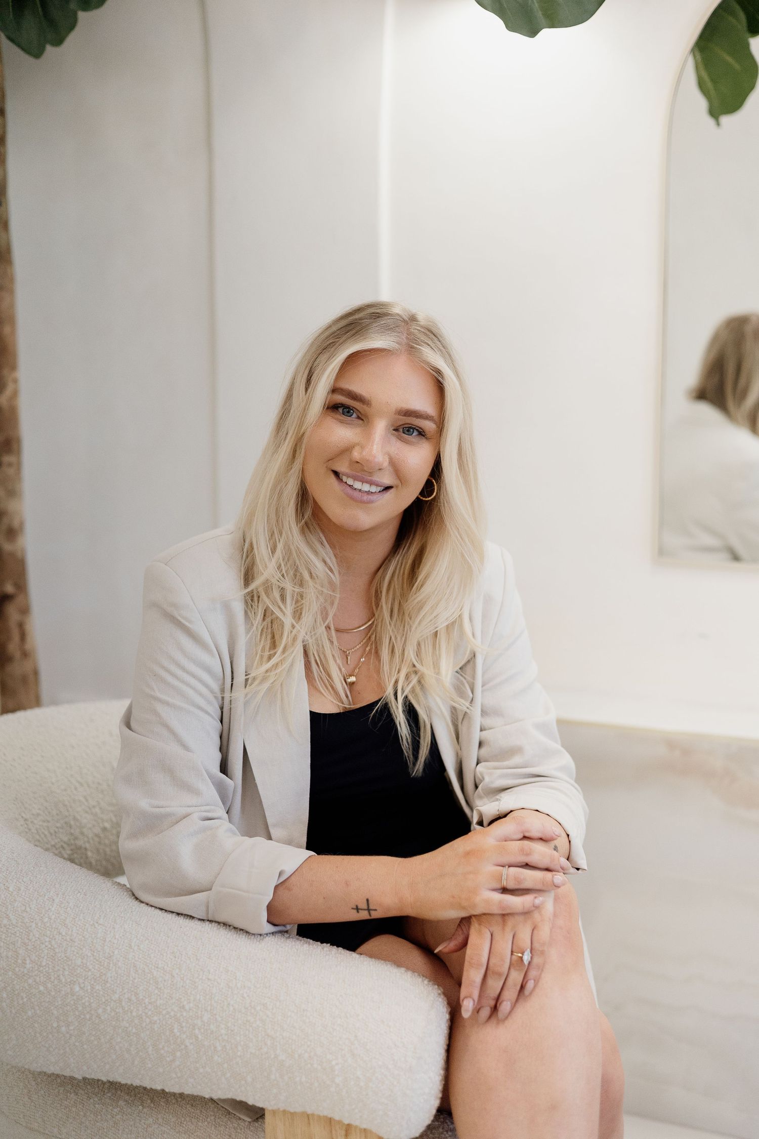 Smiling blonde woman in white blazer sitting in bright modern hair salon interior.