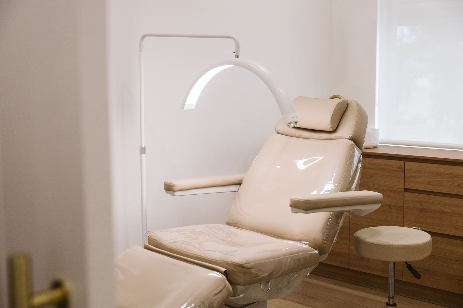 Beige leather treatment chair with overhead lamp in a modern aesthetics clinic room.